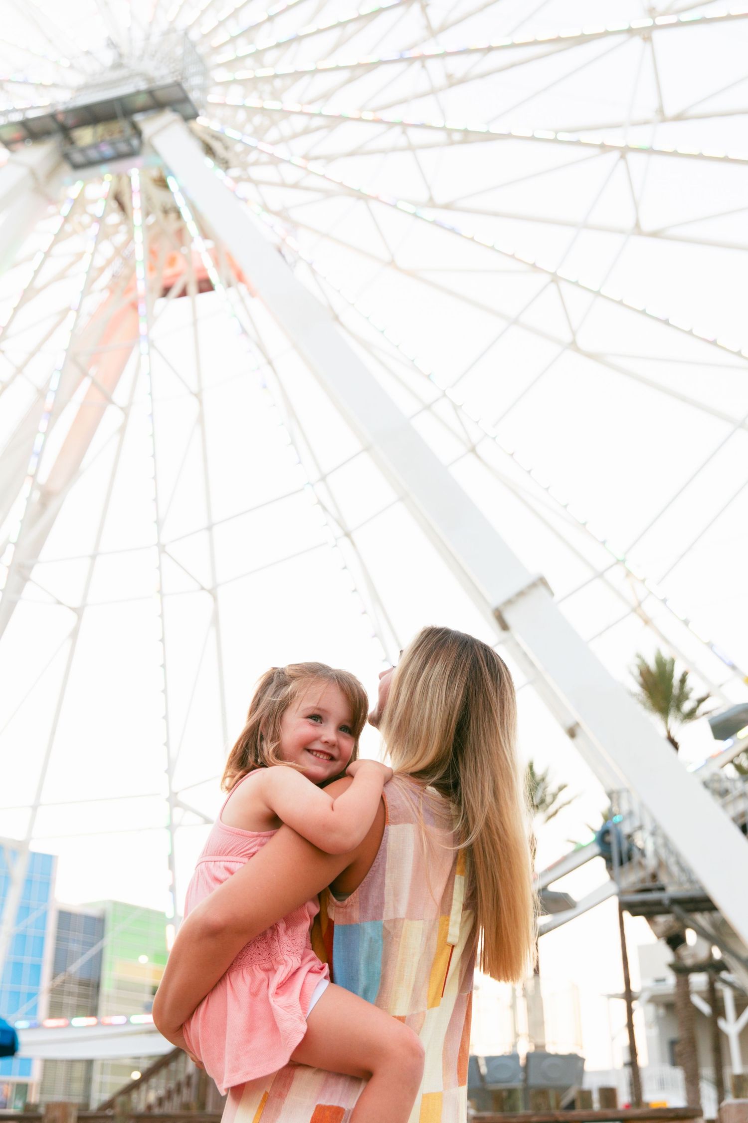 People embrace against the backdrop of a white Ferris wheel with palm trees visible at sunset.