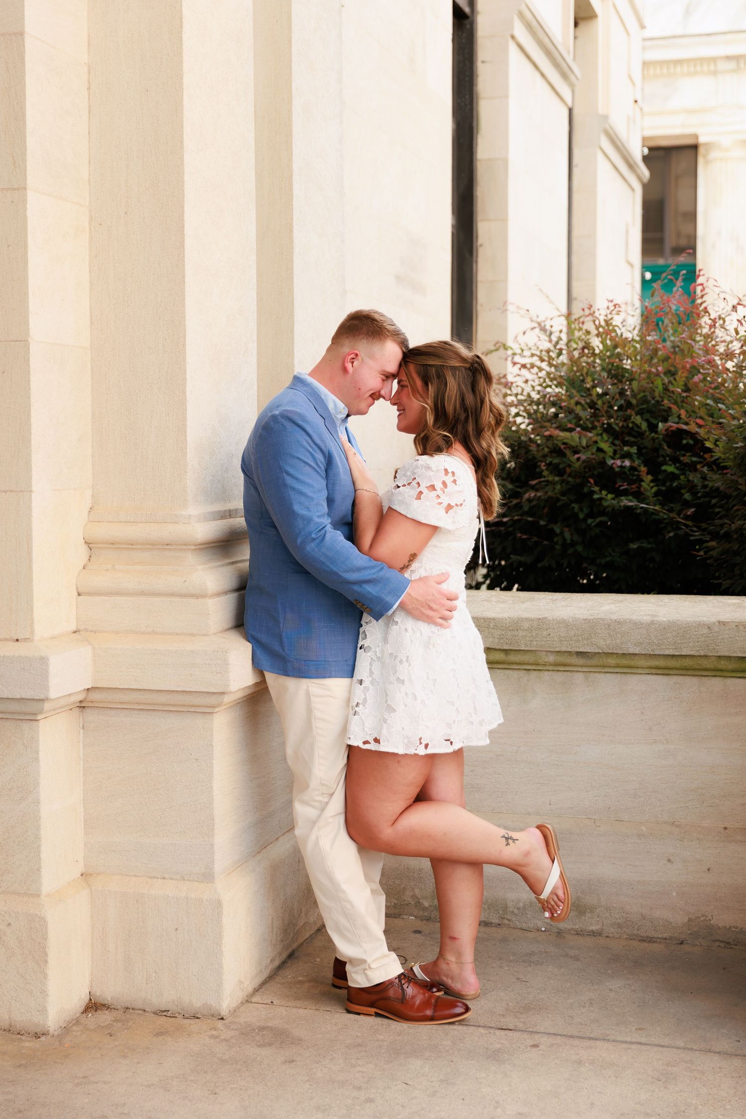 Romantic couple leaning against stone column, man in blue blazer and woman in white lace dress sharing a kiss.