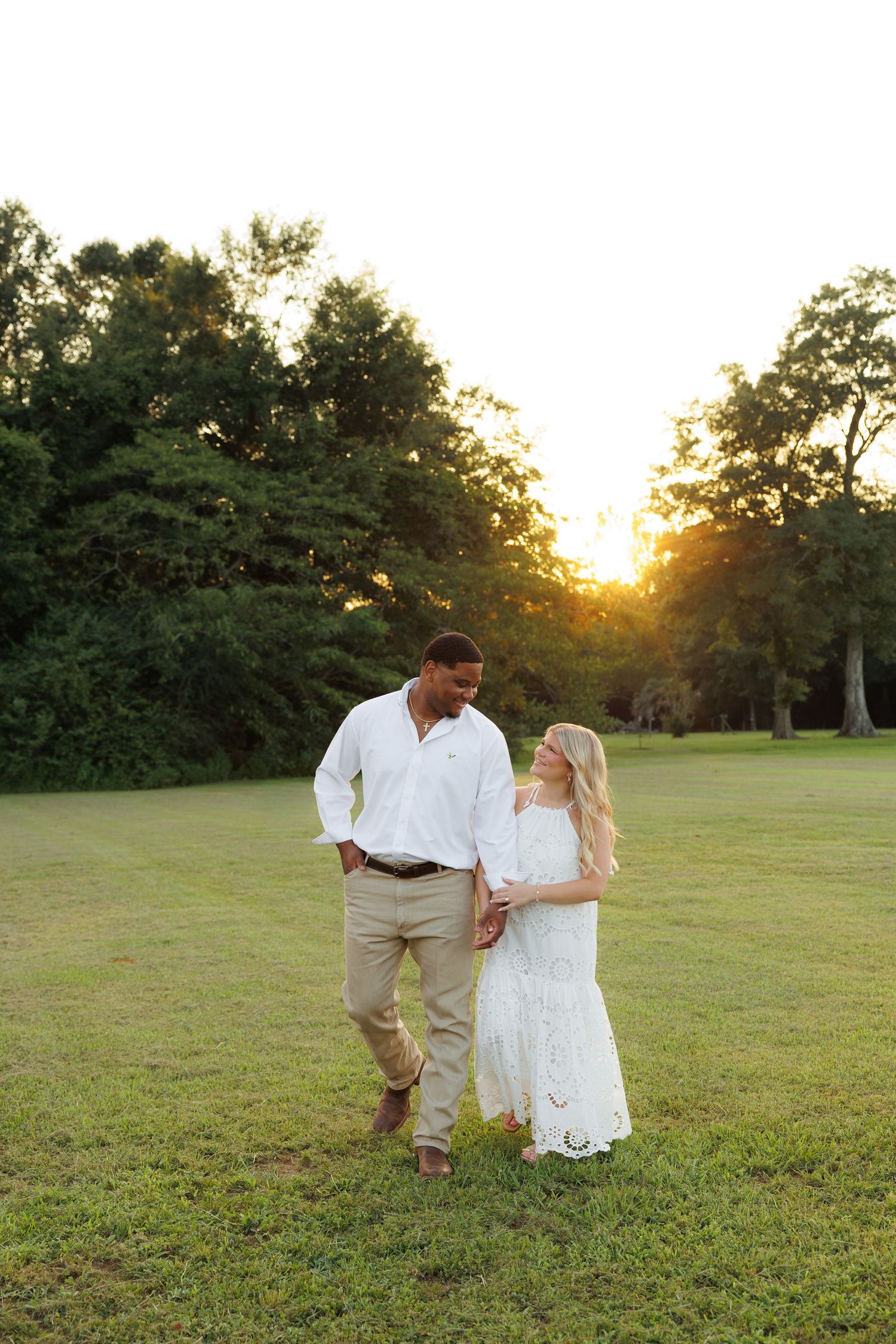 A couple walks hand in hand on a green lawn at golden hour, man in white shirt and woman in white lace dress.