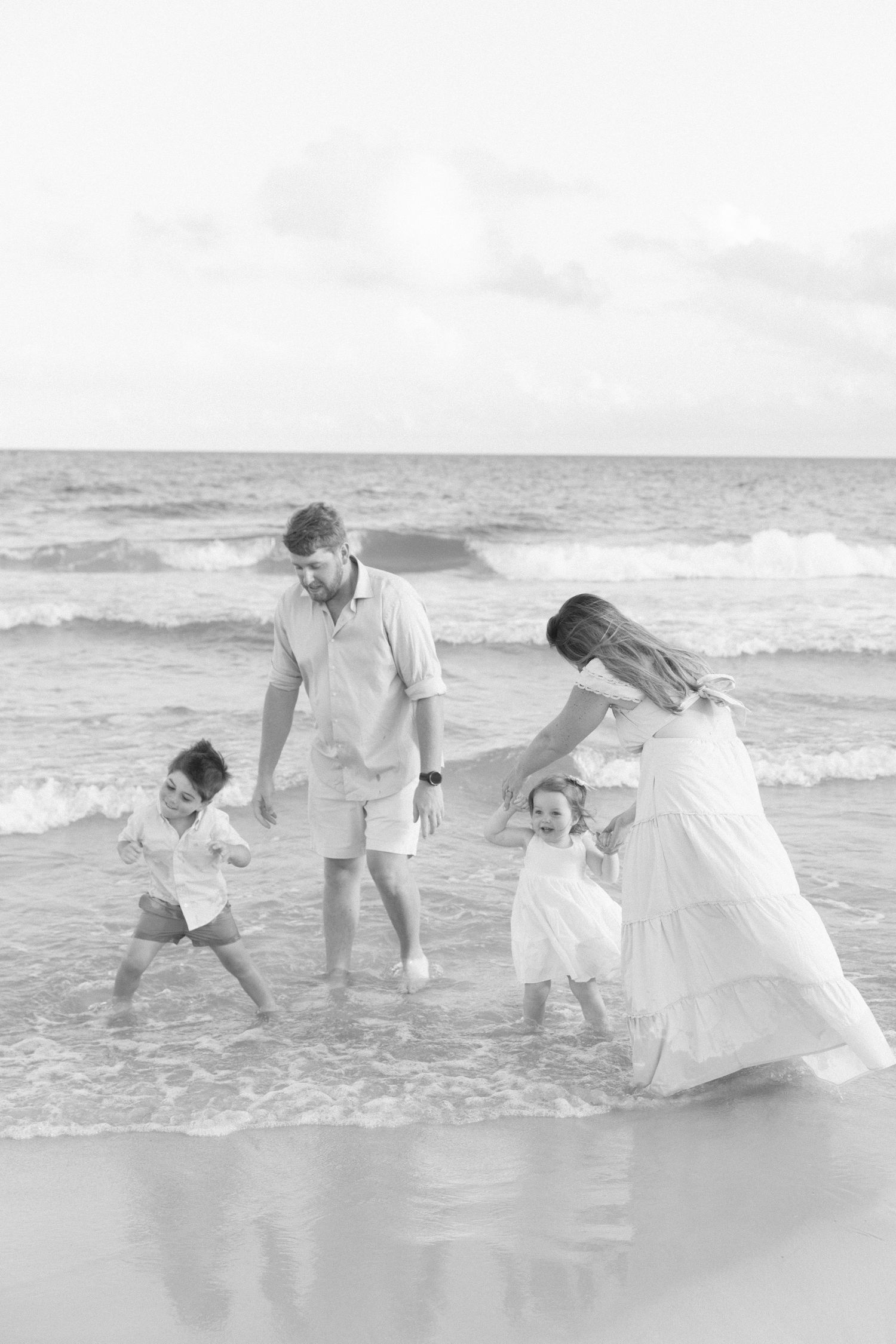 Black and white photo of a family of four playing in ocean waves on a beach.