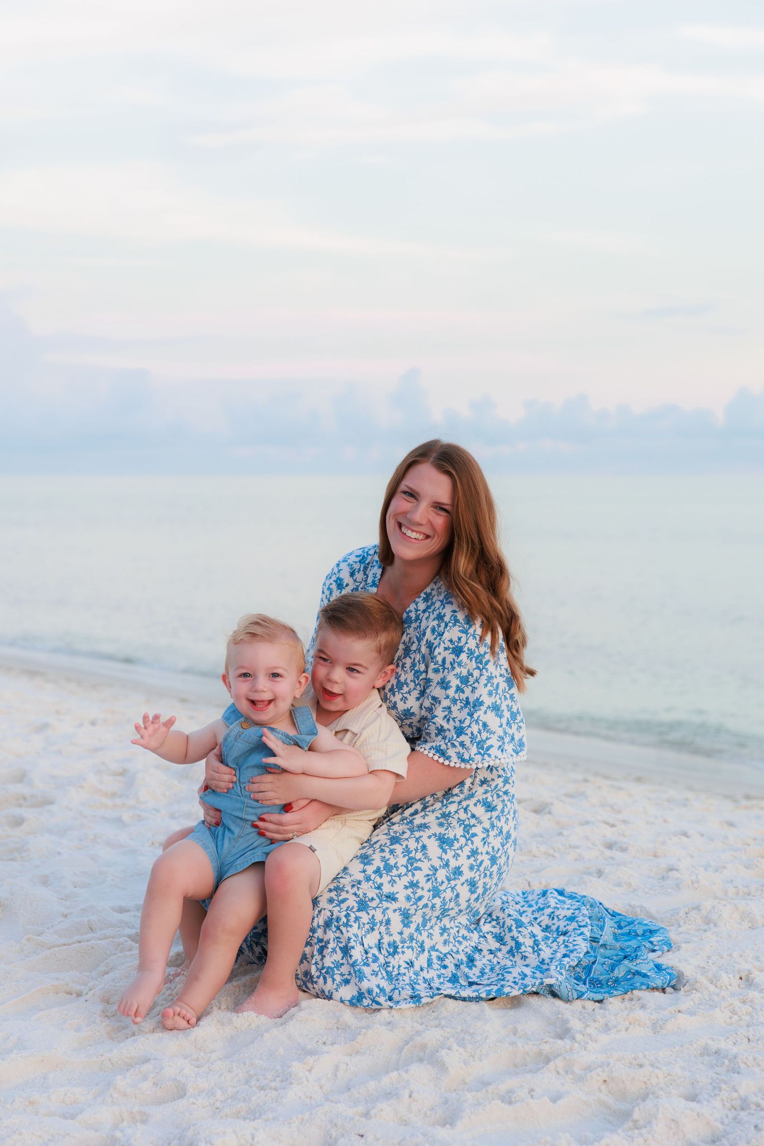A smiling mother in a blue floral dress sits on a white sand beach holding two young boys, with calm ocean waters behind them.
