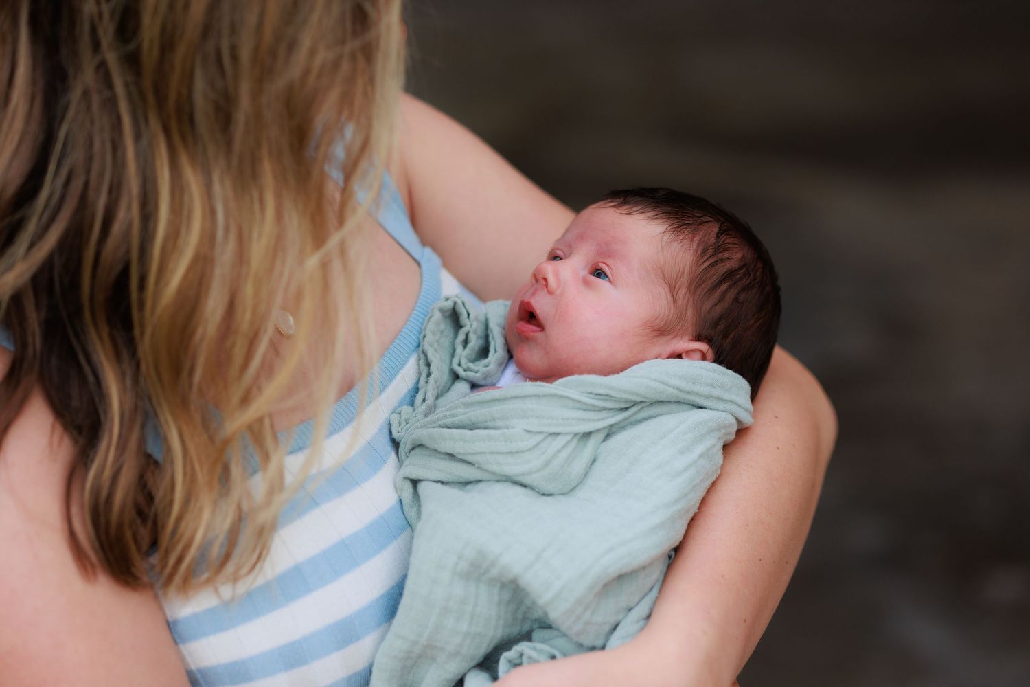A newborn baby wearing a mint green outfit is cradled in someone's arms wearing a blue and white striped shirt.