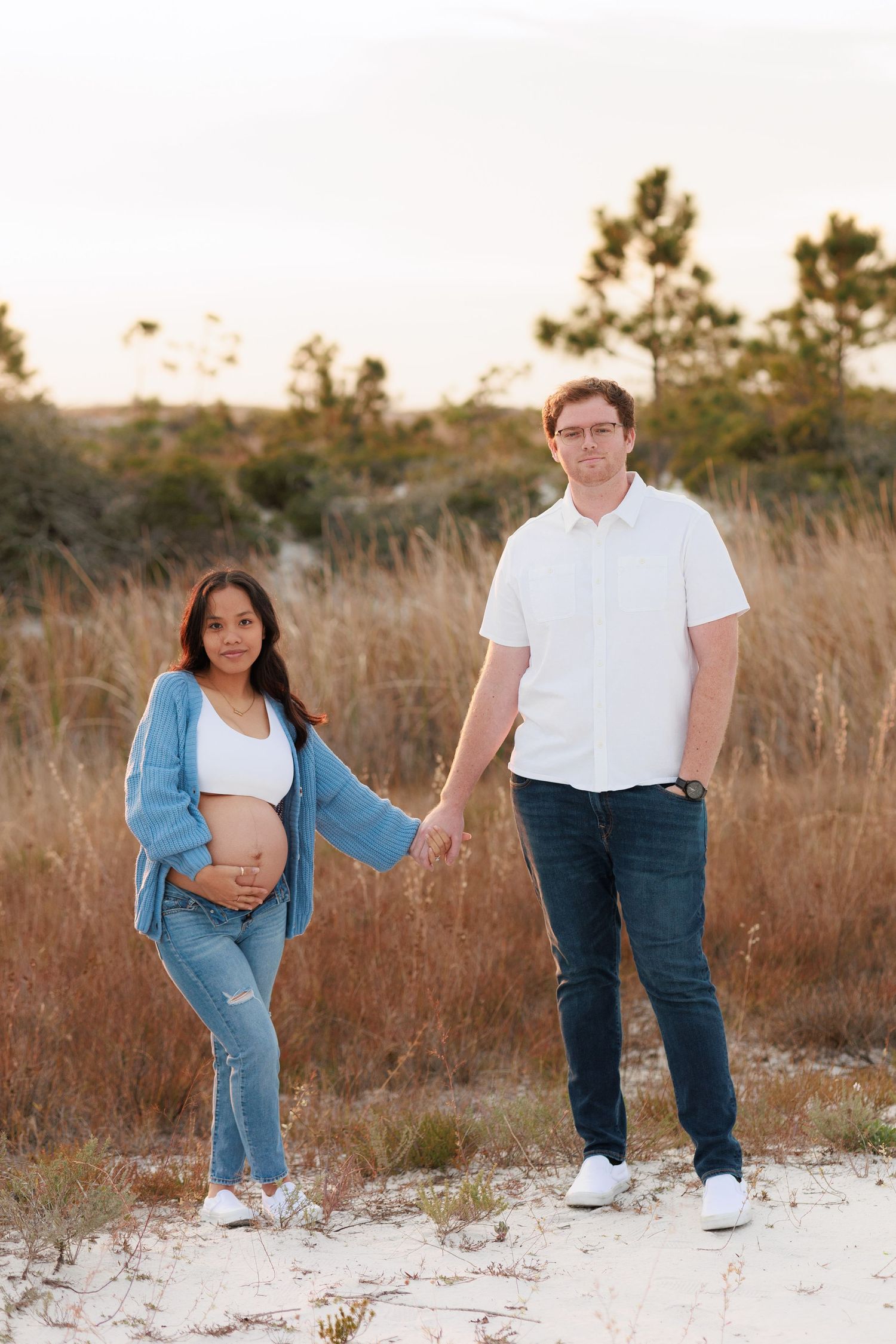 Maternity photo shoot at sunset showing couple in casual attire against tall grass and sandy backdrop.
