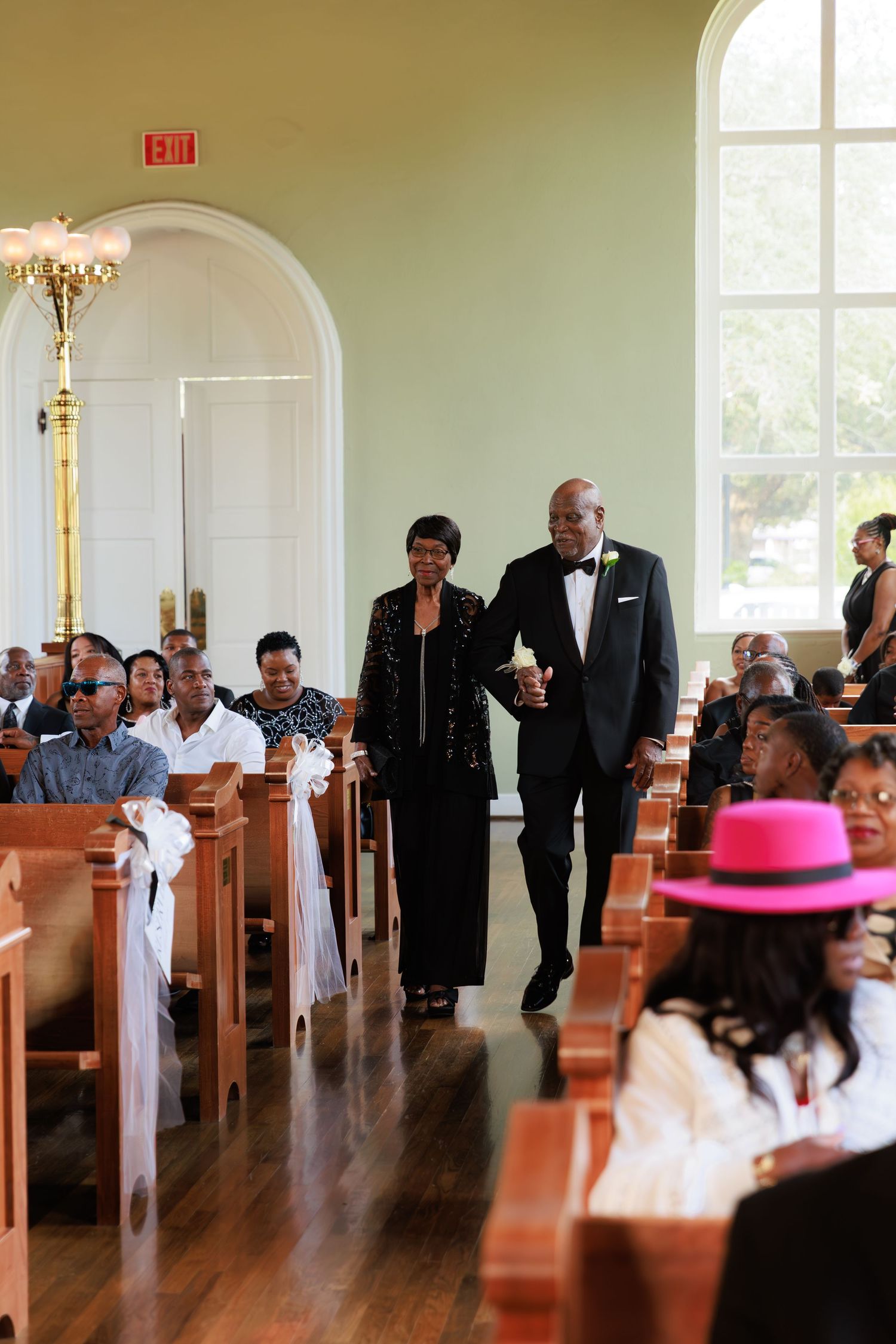 Elegant couple walks down the aisle of a traditional church with wooden pews and arched doorways during a ceremony.