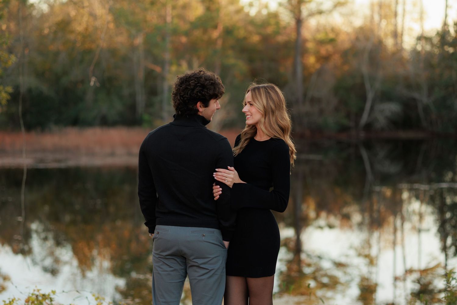 A couple in dark clothing stands together by a serene lake at sunset with autumn trees reflecting in the water.