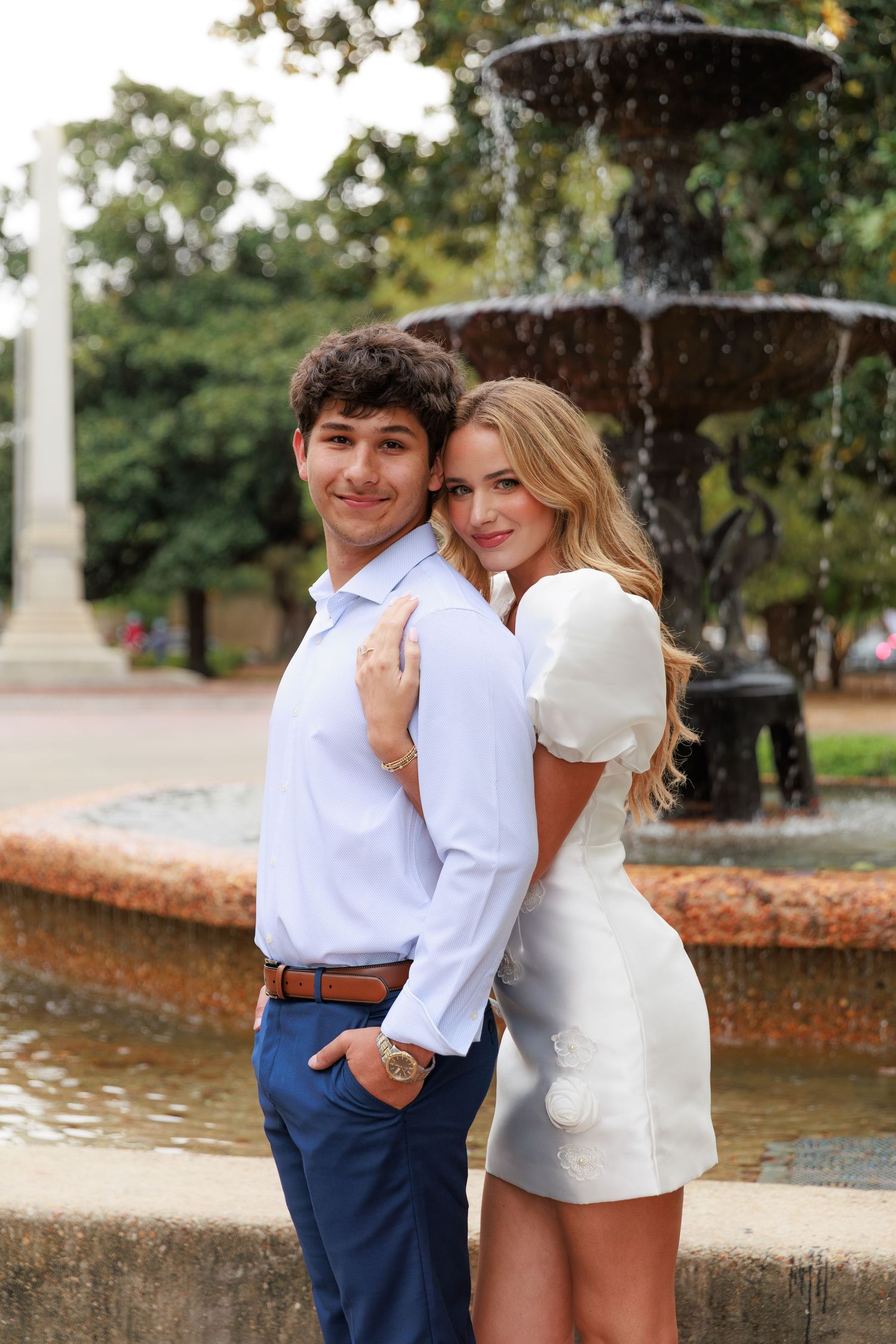 A couple poses together by a decorative fountain in a park setting, wearing coordinated white and blue outfits.