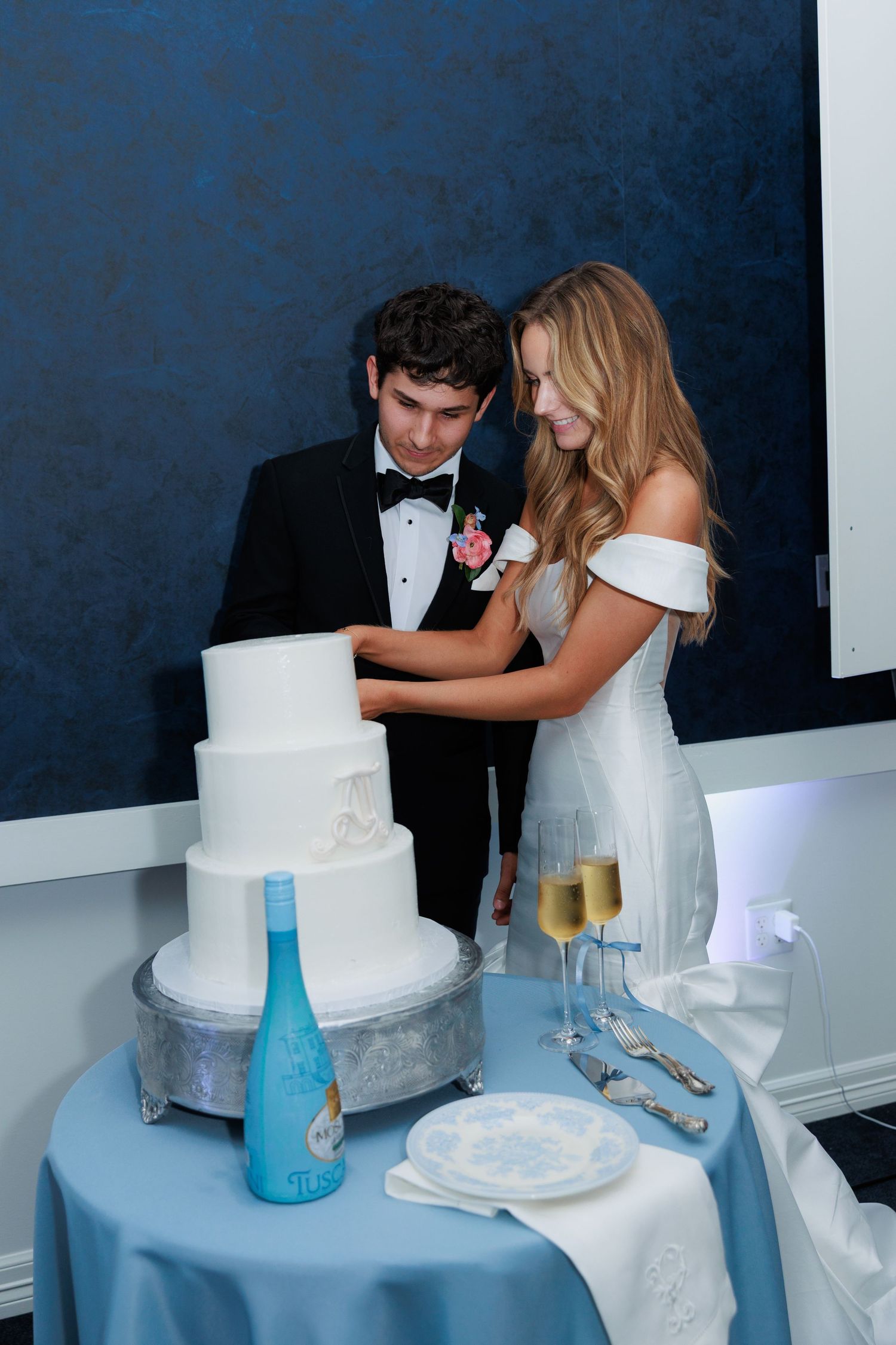 A wedding couple cutting their elegant white tiered cake next to blue glass vases on a table.