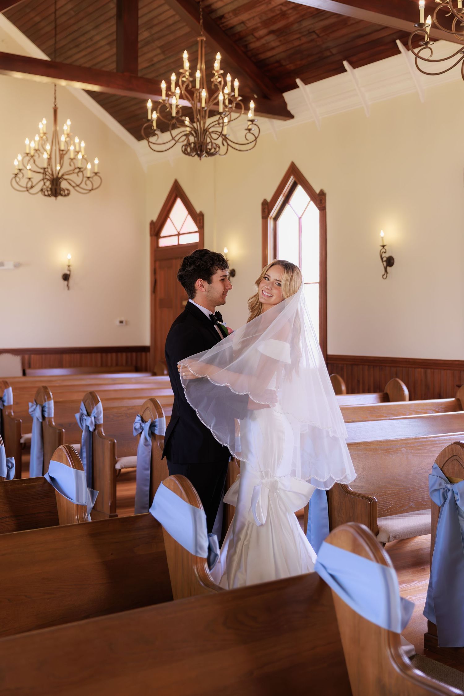 Elegant chapel interior with chandeliers and blue ribbon pew decor during a wedding ceremony.