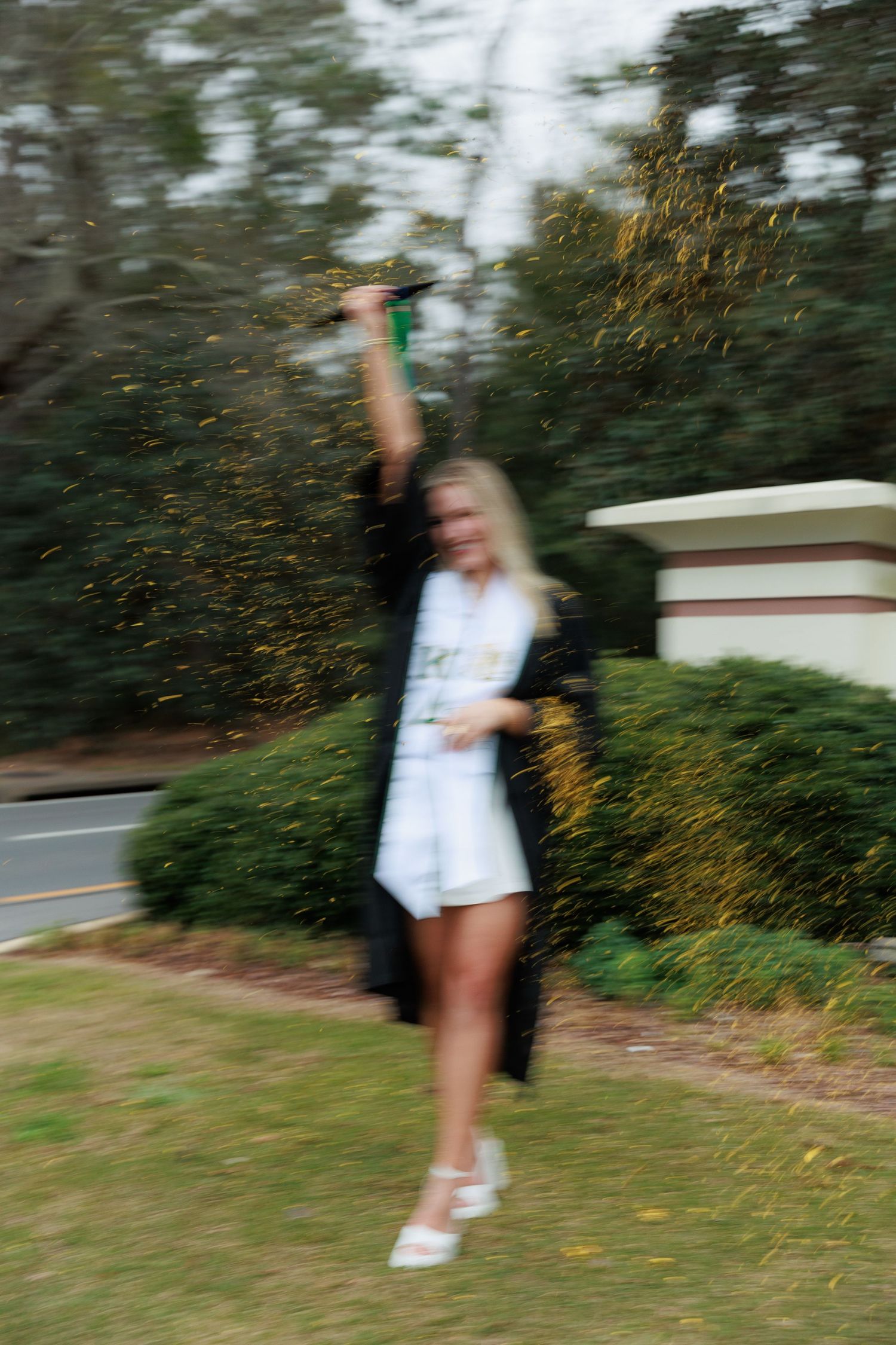 Motion blur photo of blonde graduate in black gown celebrating with confetti outdoors on campus.