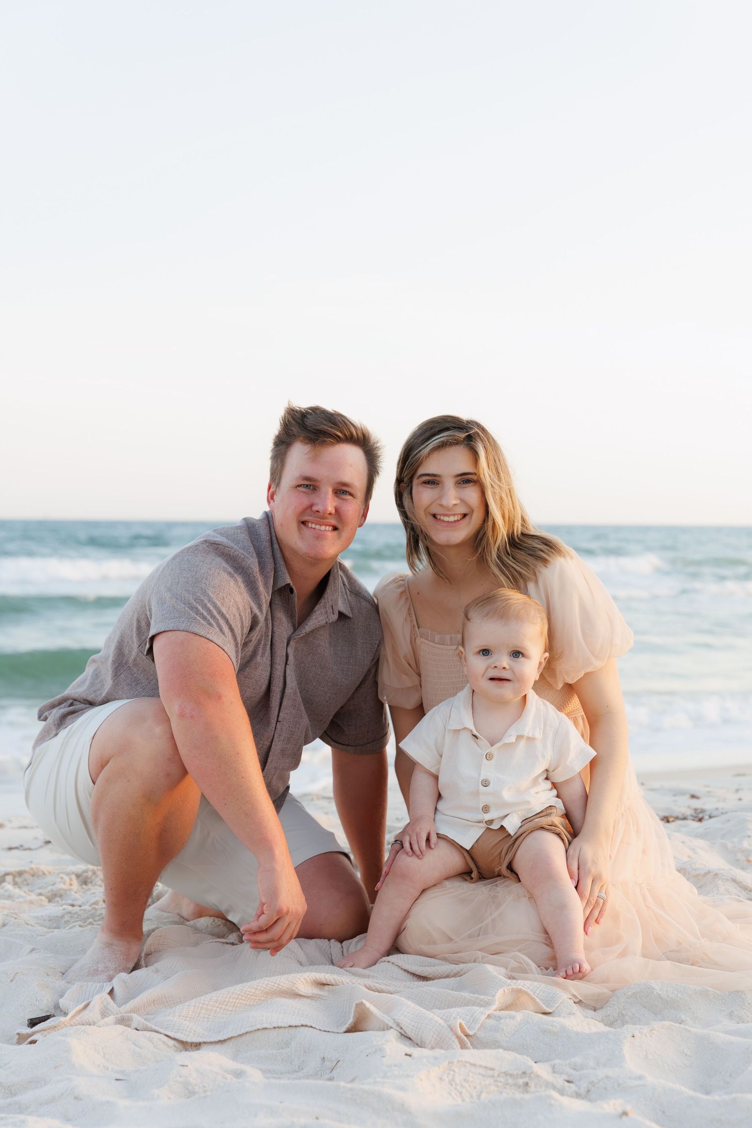 Happy family with a baby boy posing on a sandy beach at sunset, smiling at the camera.