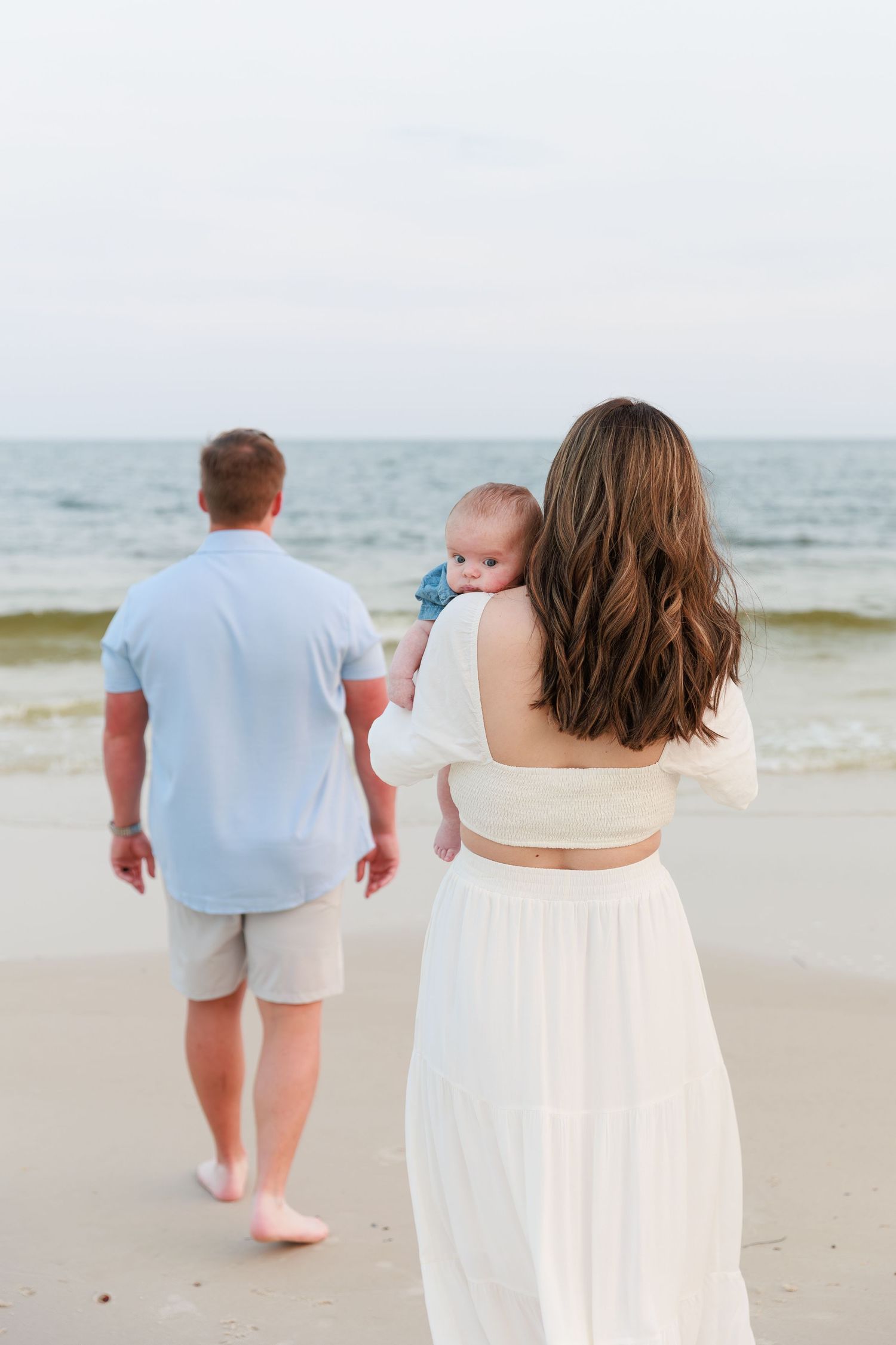A young family with a baby walks along a sandy beach, with the ocean in the background.