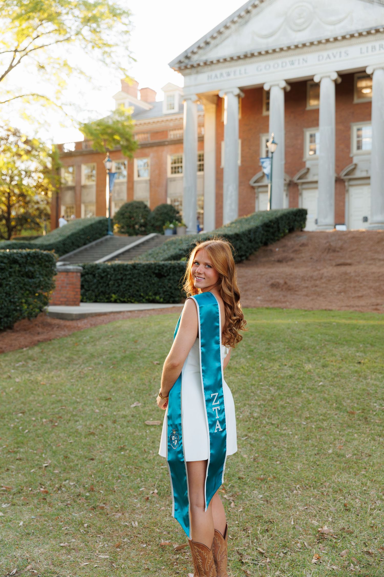 Young woman in white dress with teal graduation sash poses on college campus lawn before brick building with columns.