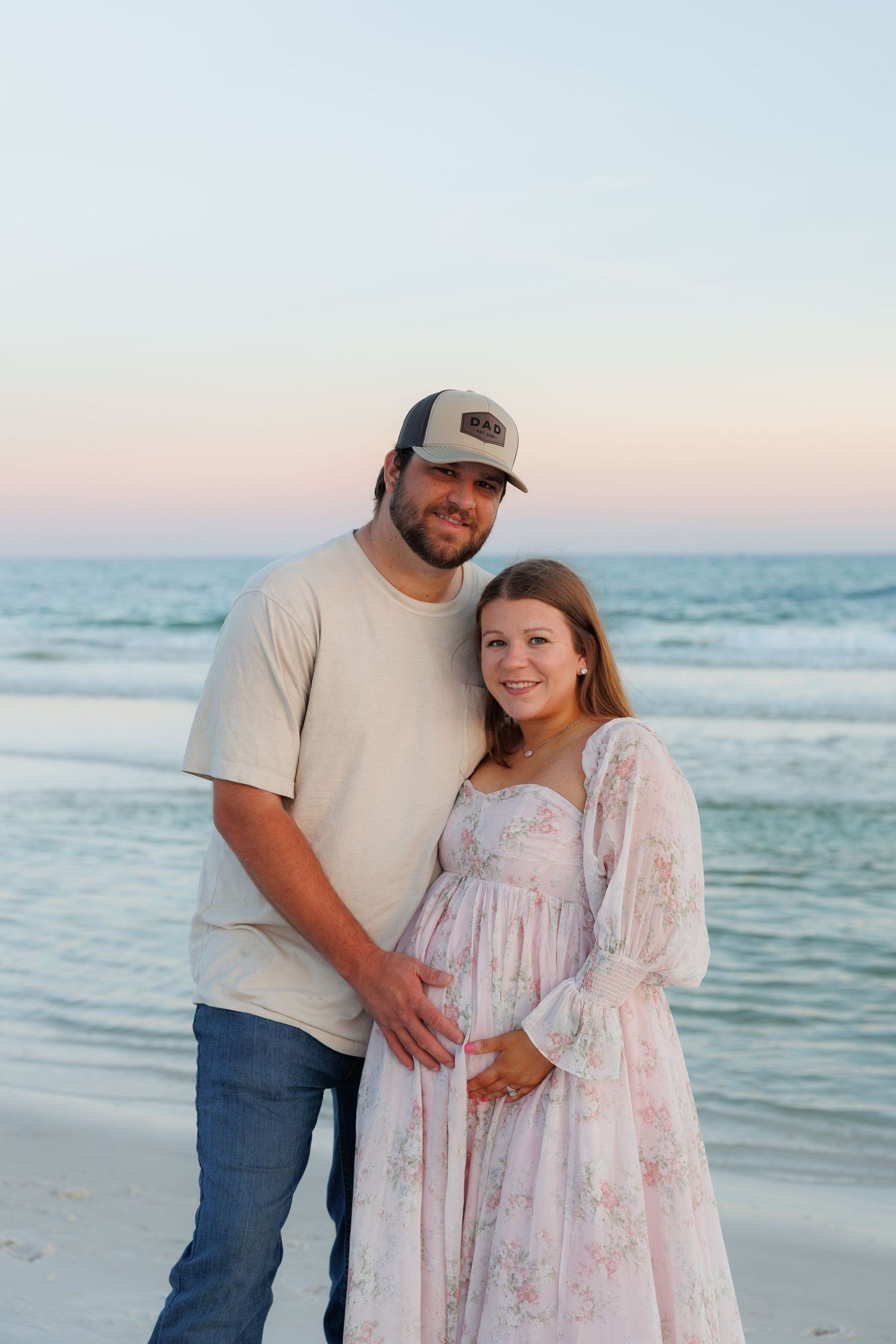 Pregnant couple posing together on a beach at sunset, woman in floral maternity dress, man in cap and jeans.