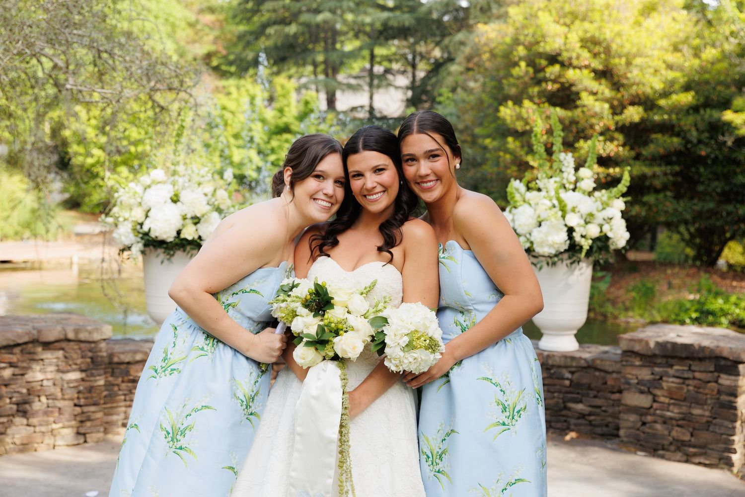 Bride in white gown poses with two bridesmaids in light blue floral dresses, holding white bouquets outdoors.