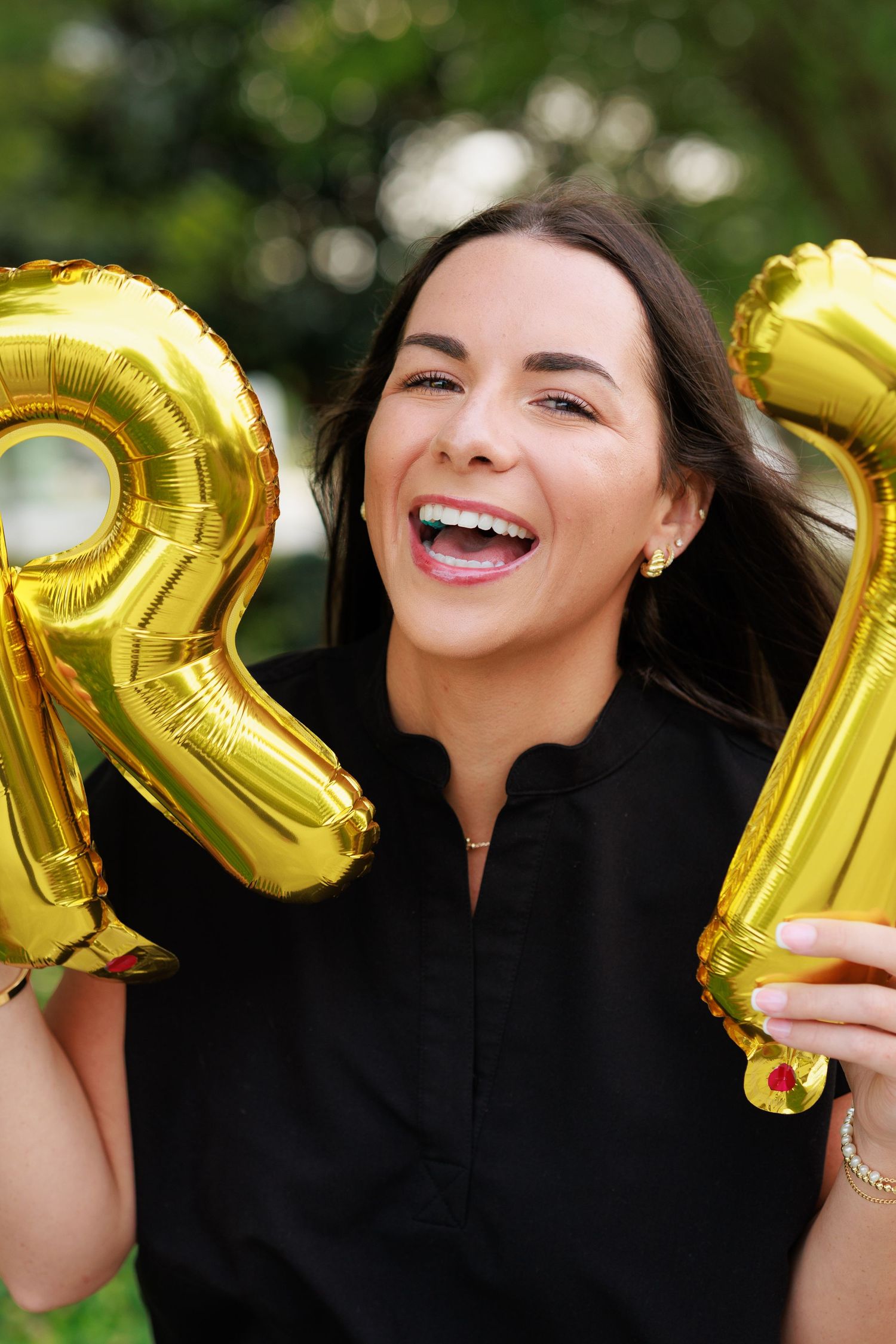 A laughing woman in black dress holds gold letter R and I balloons outdoors.