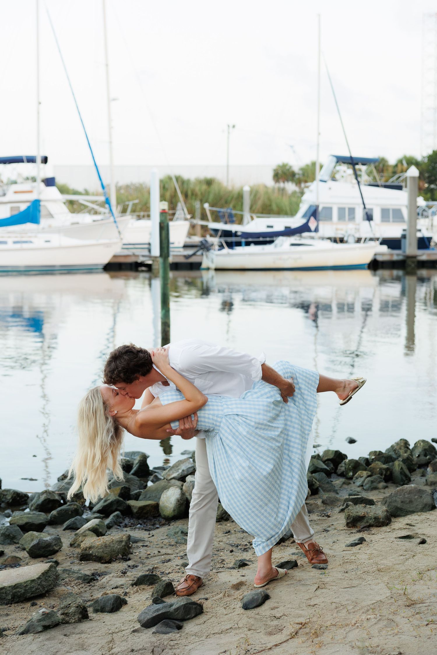 Couple sharing a romantic dip kiss on a sandy marina shore with sailboats and calm water in the background.