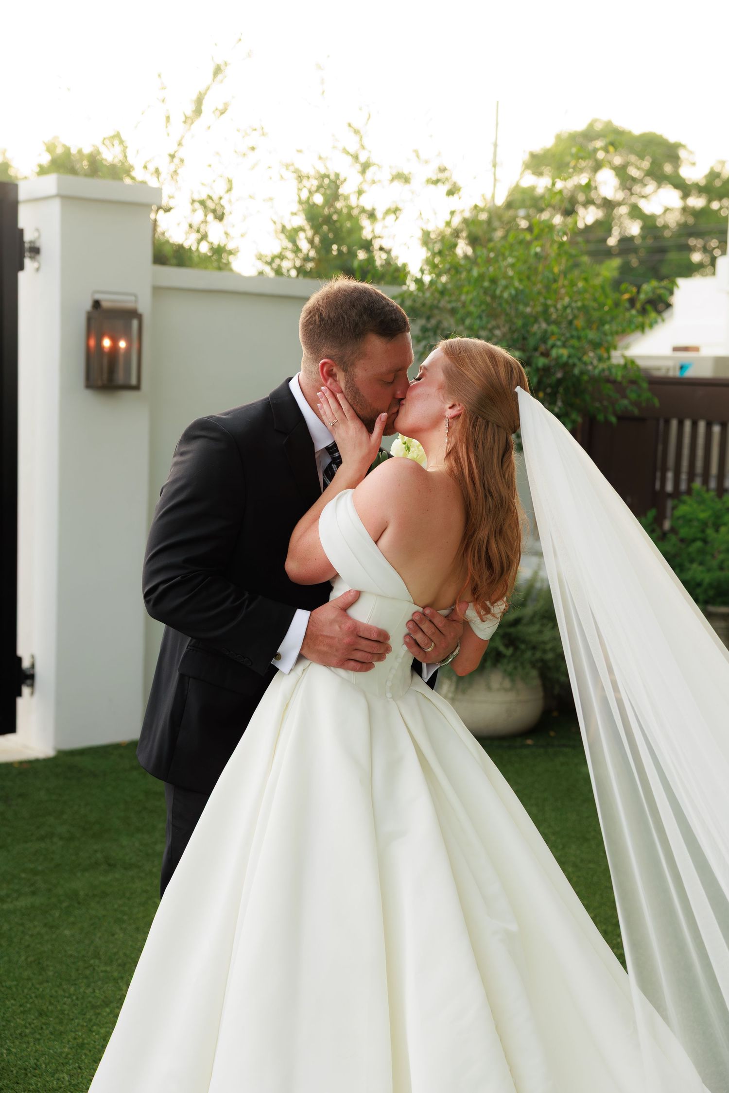 Newlyweds share a romantic kiss outdoors, bride in off-shoulder gown with flowing veil in garden setting.