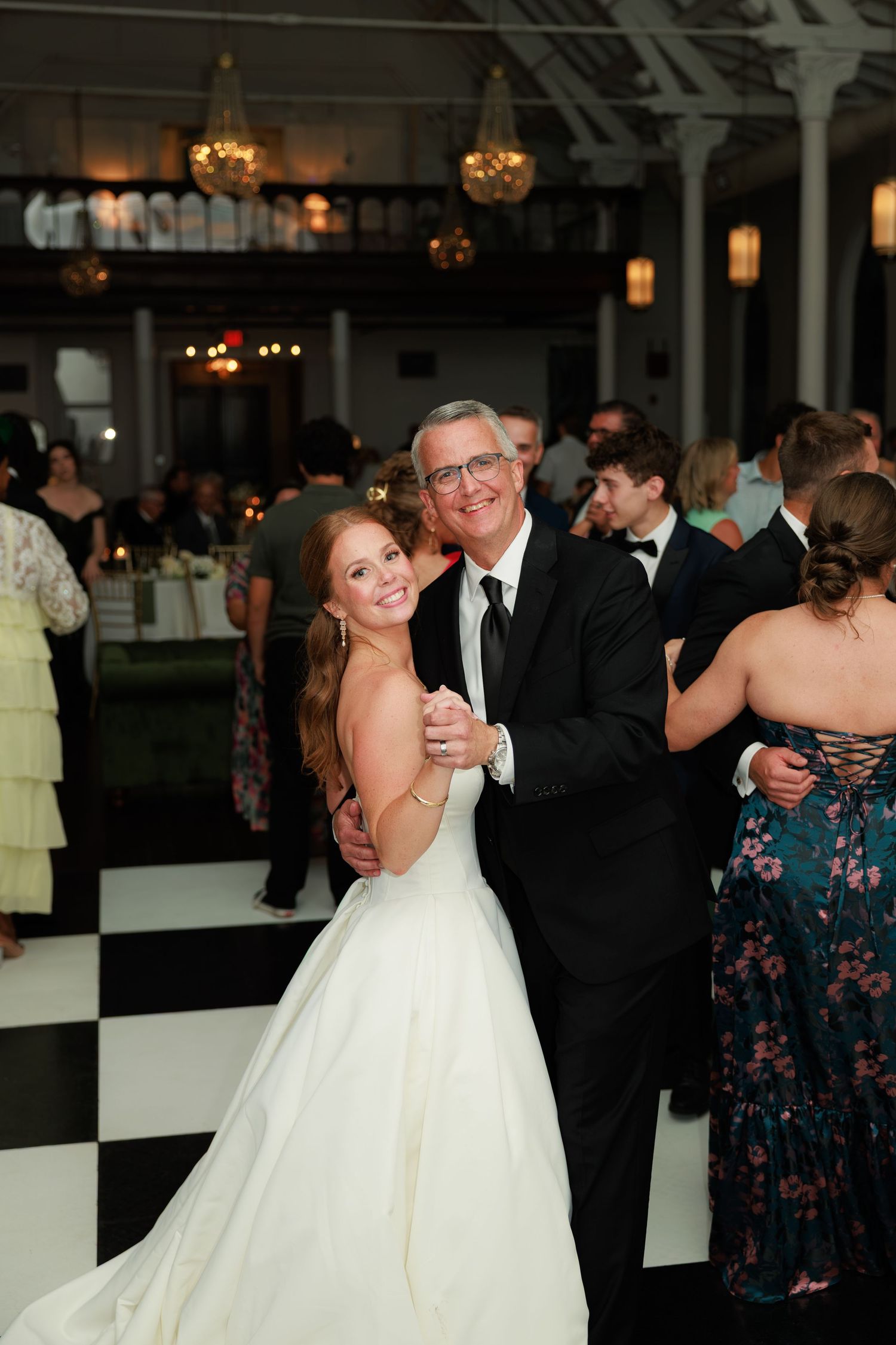 Smiling bride dances with her father on a classic black and white checkered dance floor at wedding reception.
