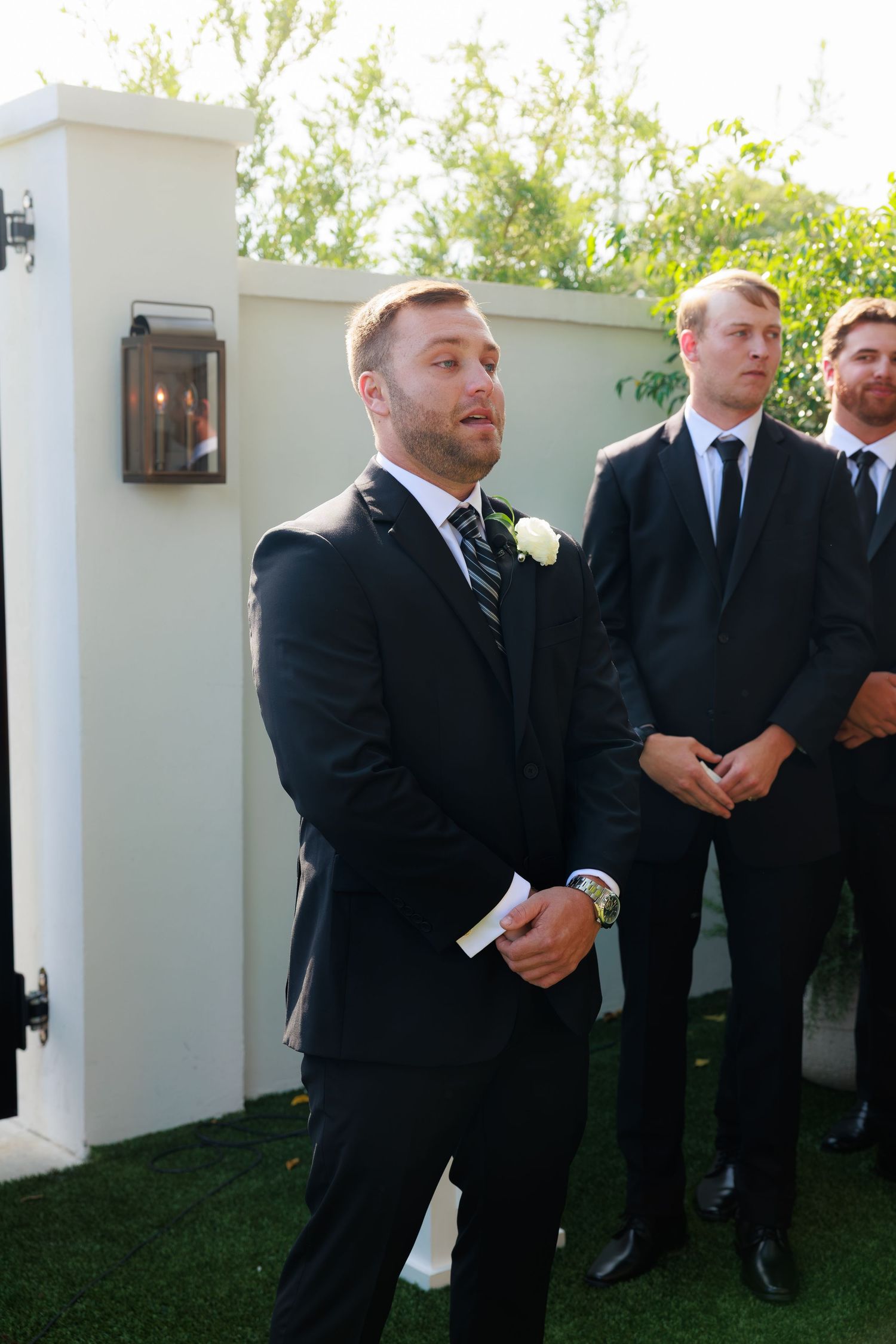 Groom in black suit with white boutonniere stands at outdoor wedding ceremony with groomsmen behind him.