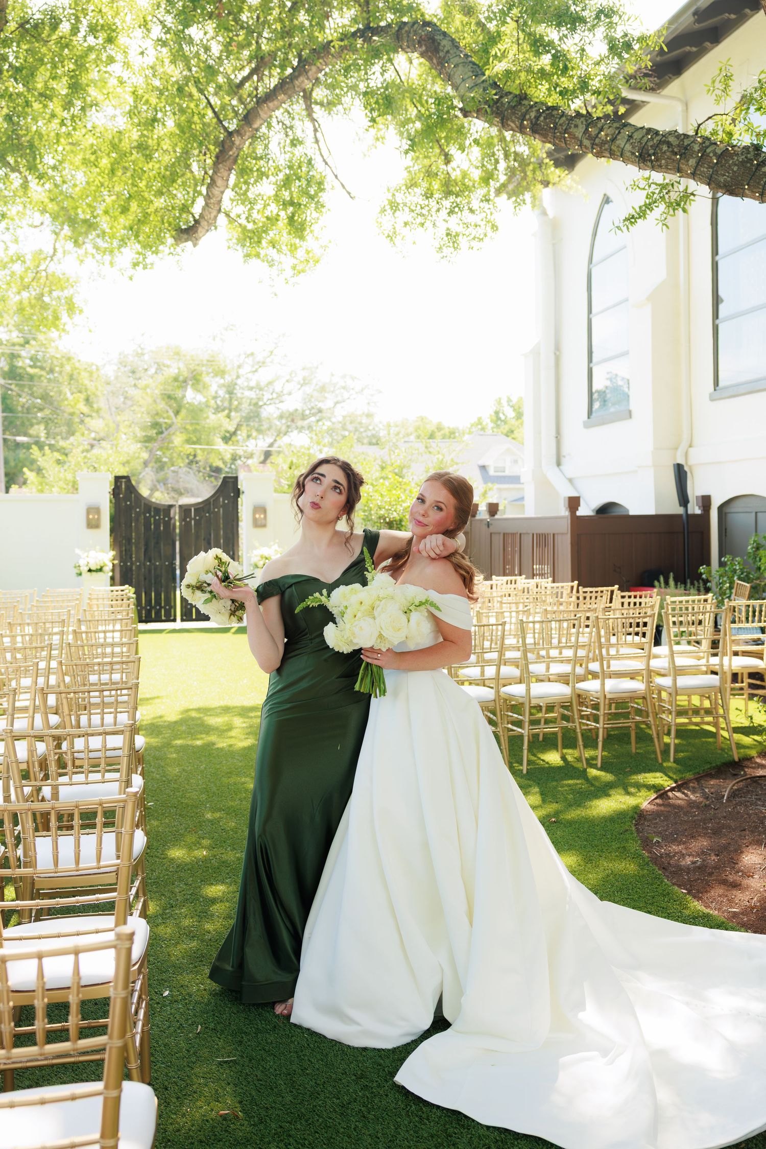 Bride in white gown and bridesmaid in green dress holding bouquets at outdoor wedding ceremony with gold chairs.