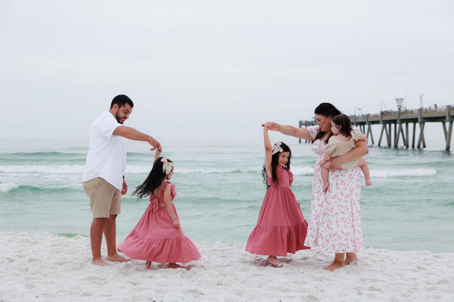Family dancing on a sandy beach near a pier, with two girls in pink maxi dresses and a woman holding a baby.