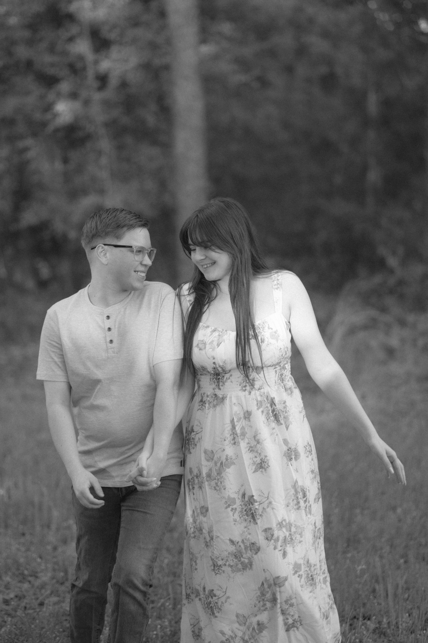 Black and white photo of a young couple smiling at each other while standing in a lush green forest setting.