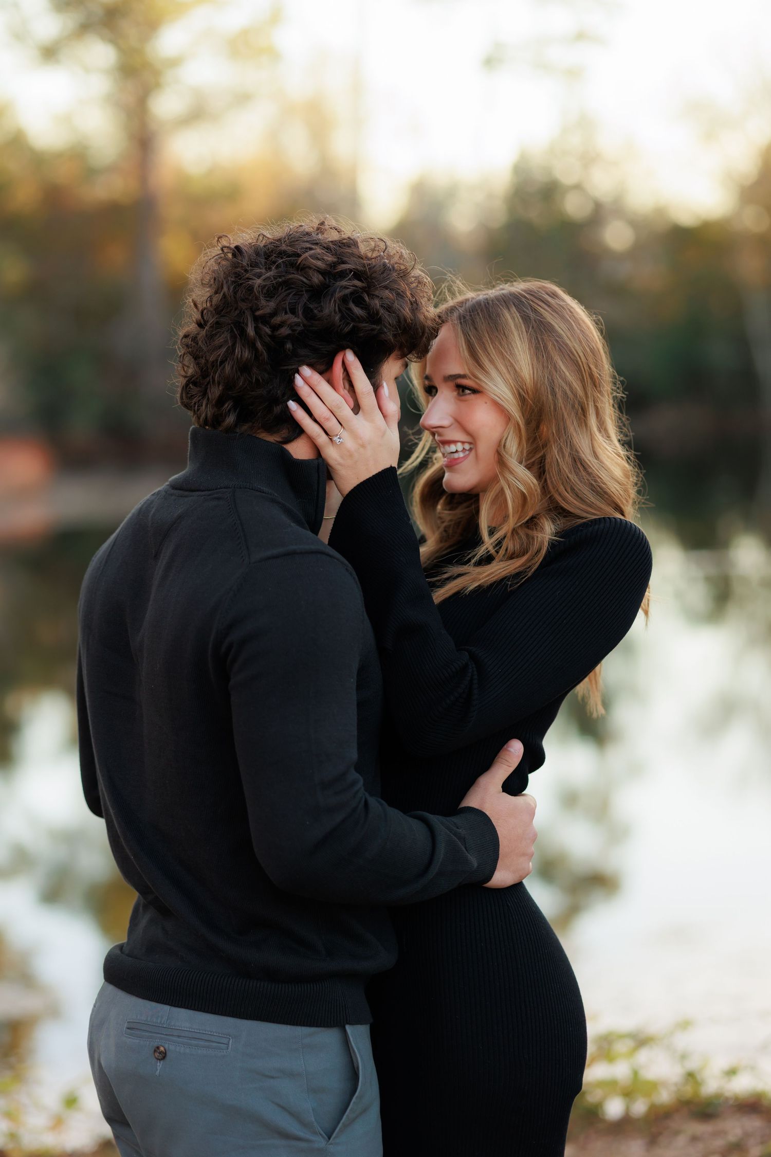 A couple laughs together during a romantic engagement photo session by a lake at sunset.