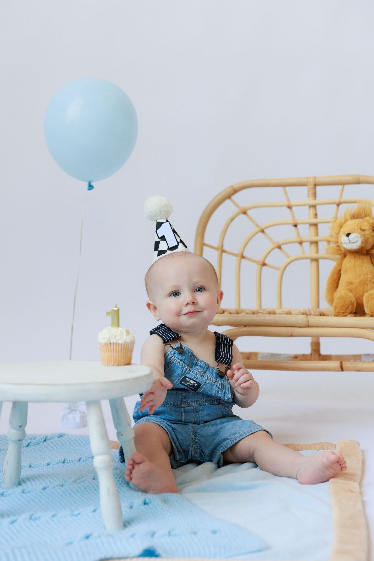 Baby in denim overalls sits on blue blanket with balloon and cake during first birthday photo session.