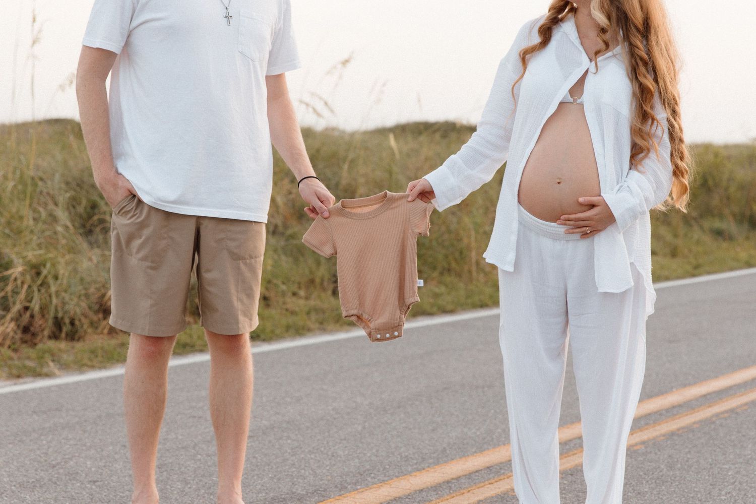 A couple in light colored clothing embraces while walking on a paved path through a grassy landscape at sunset.