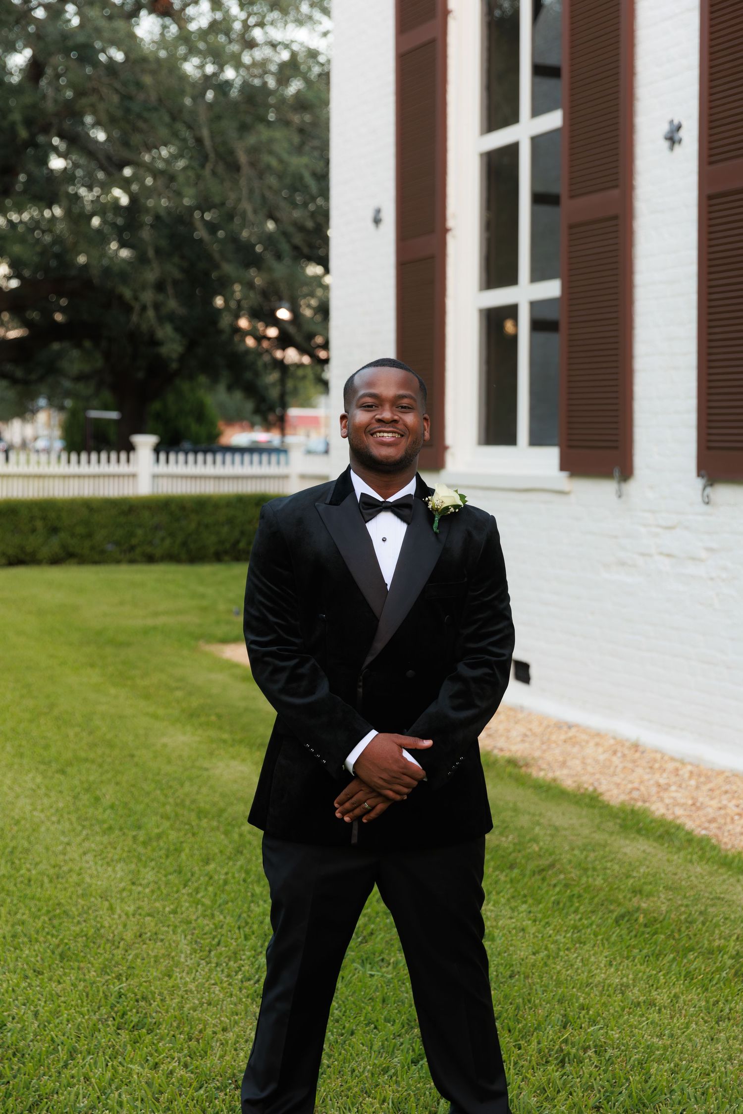 A groom in classic black tuxedo poses on church grounds with a boutonniere.