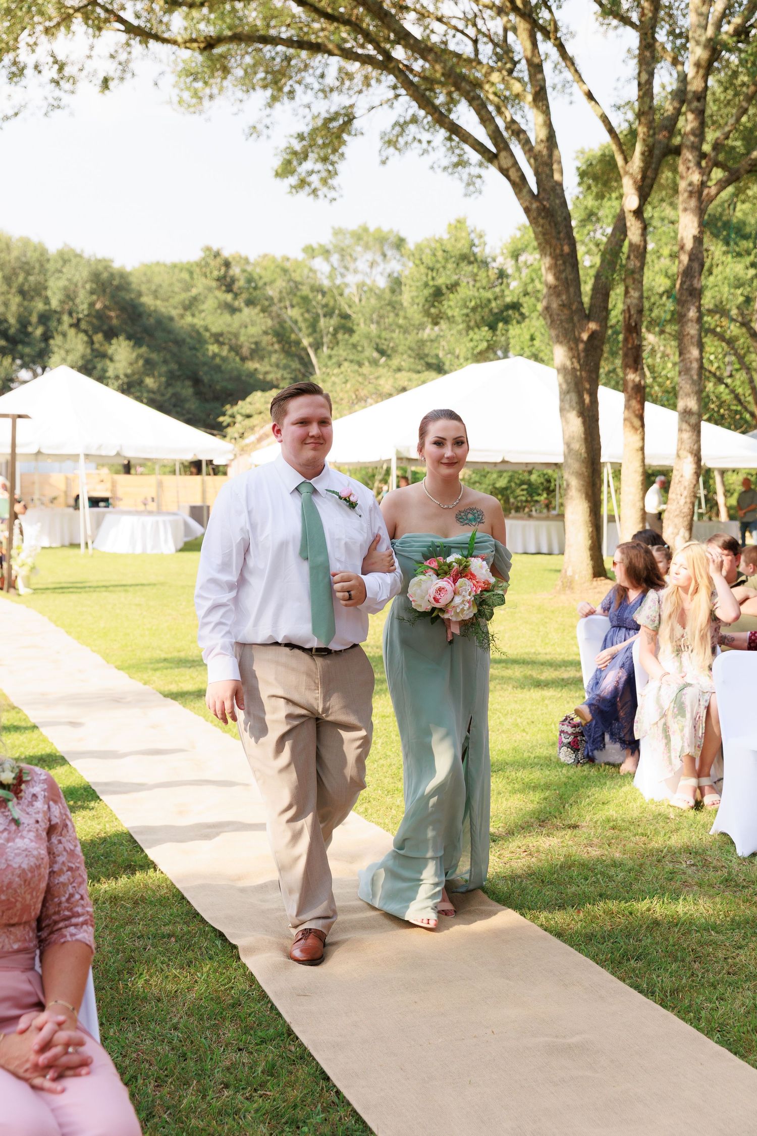 Wedding party members walk down a paved outdoor pathway lined with trees during a sunny outdoor ceremony.