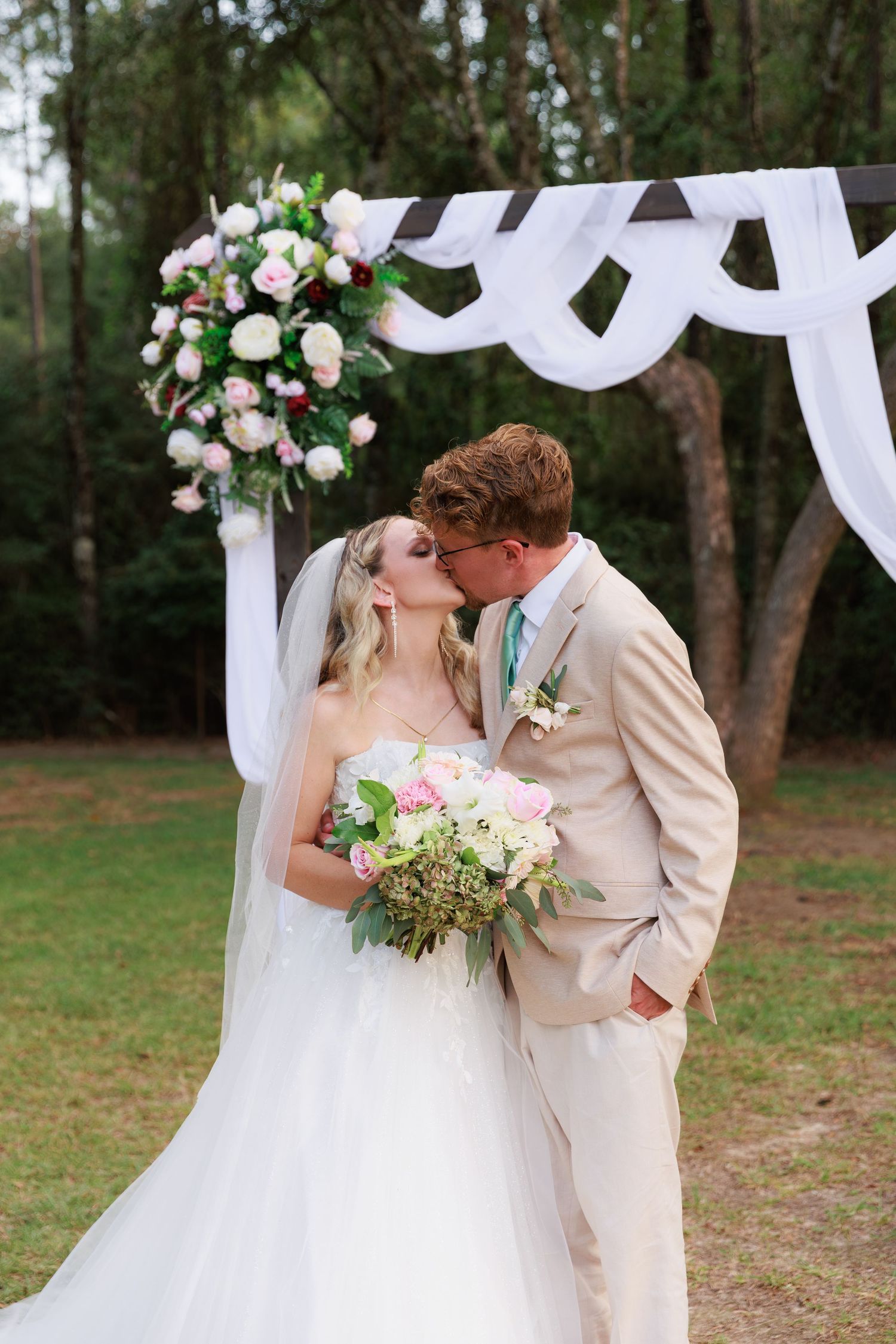 A romantic outdoor wedding ceremony under a white draped arch decorated with pink and white floral arrangements.