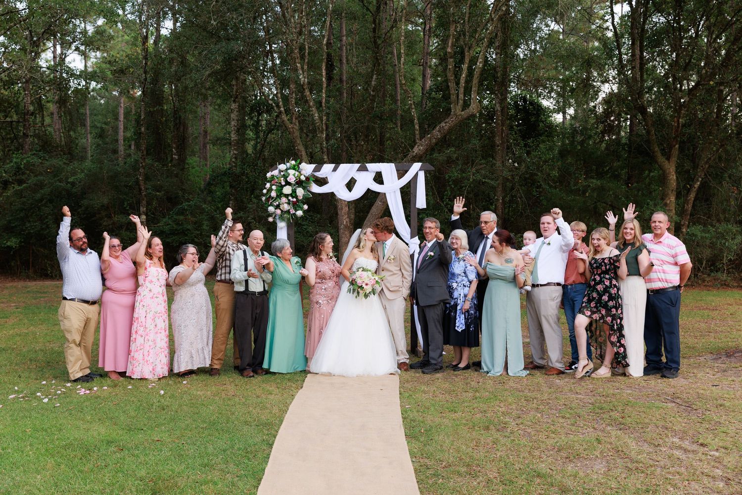Wedding party members in pastel dresses and suits pose together on a grassy lawn during an outdoor celebration.