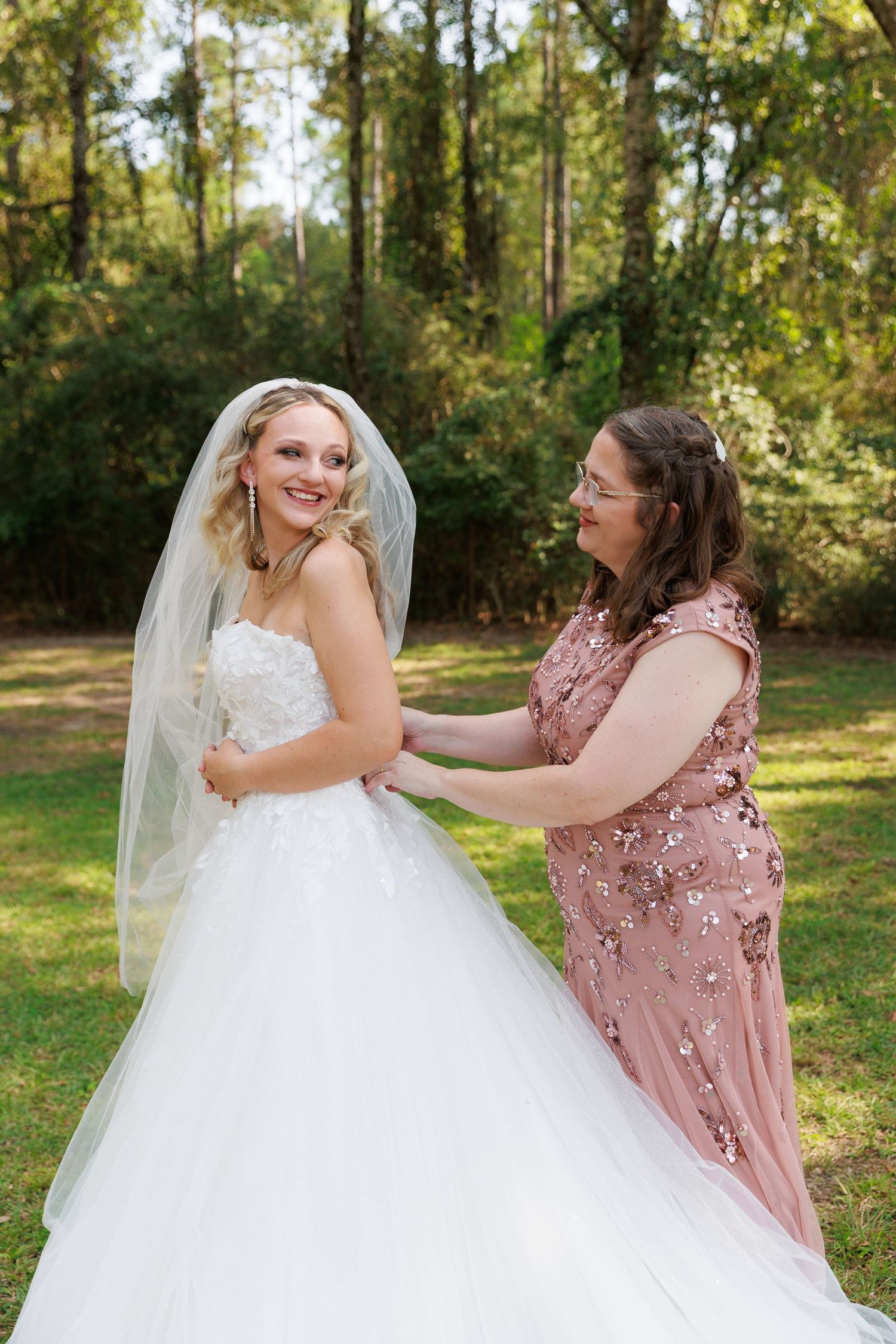 A bride in a white ballgown twirls with someone wearing a dusty rose dress in a sunlit wooded outdoor setting.
