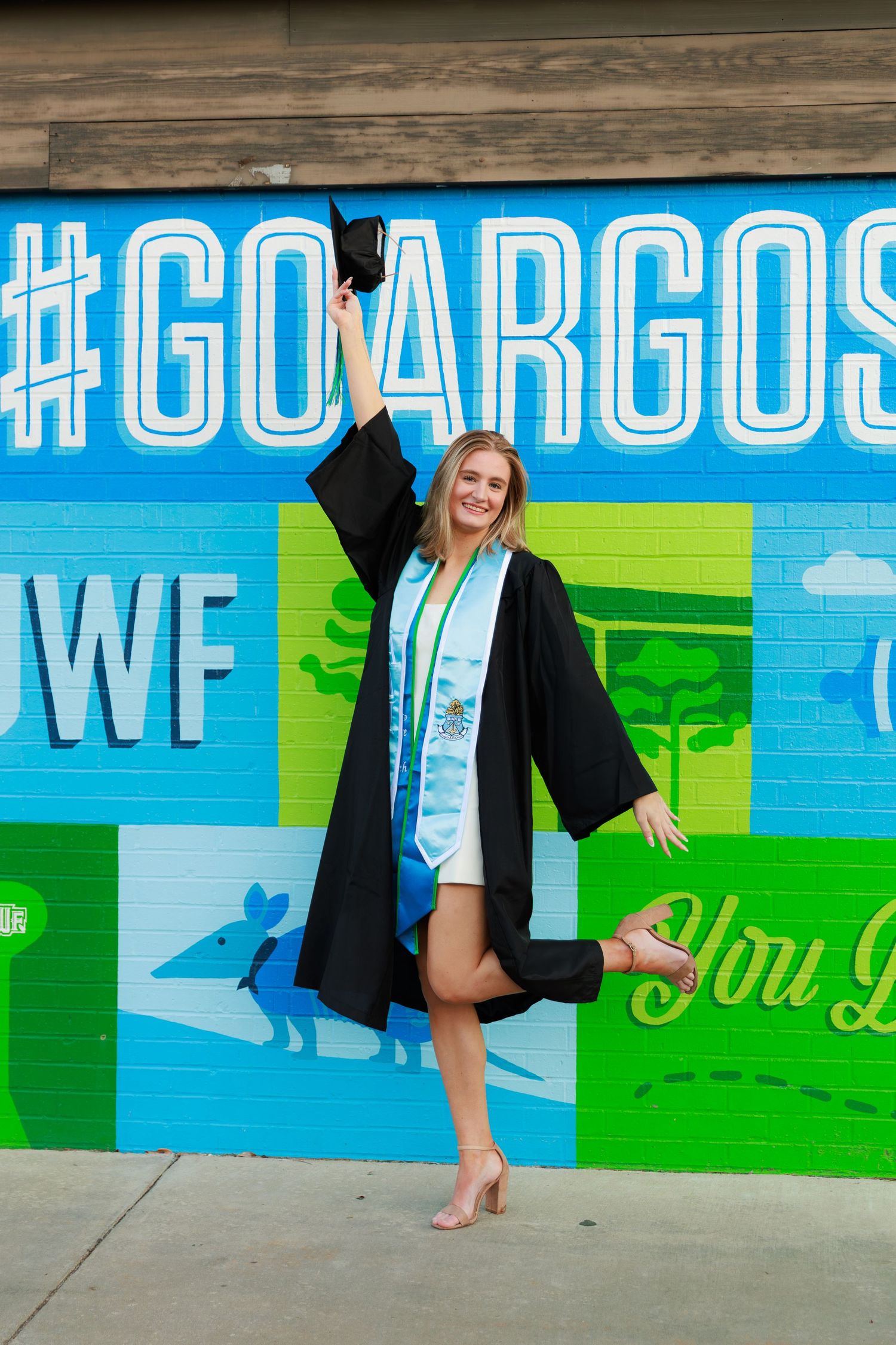 A person wearing a black graduation gown poses playfully against a vibrant blue and green painted wall mural.