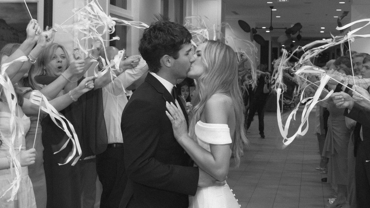 A wedding celebration captured in black and white with streamers and guests lining a hallway as the couple shares a kiss.