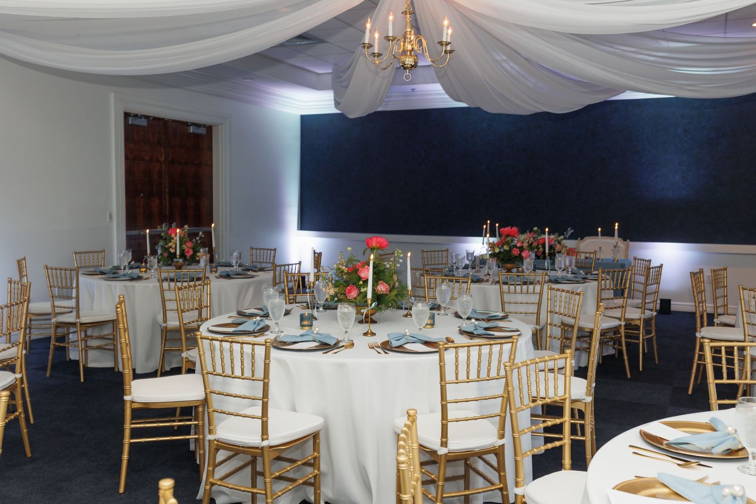 An elegant wedding reception setup with round tables, gold chiavari chairs, and white draping under a crystal chandelier.