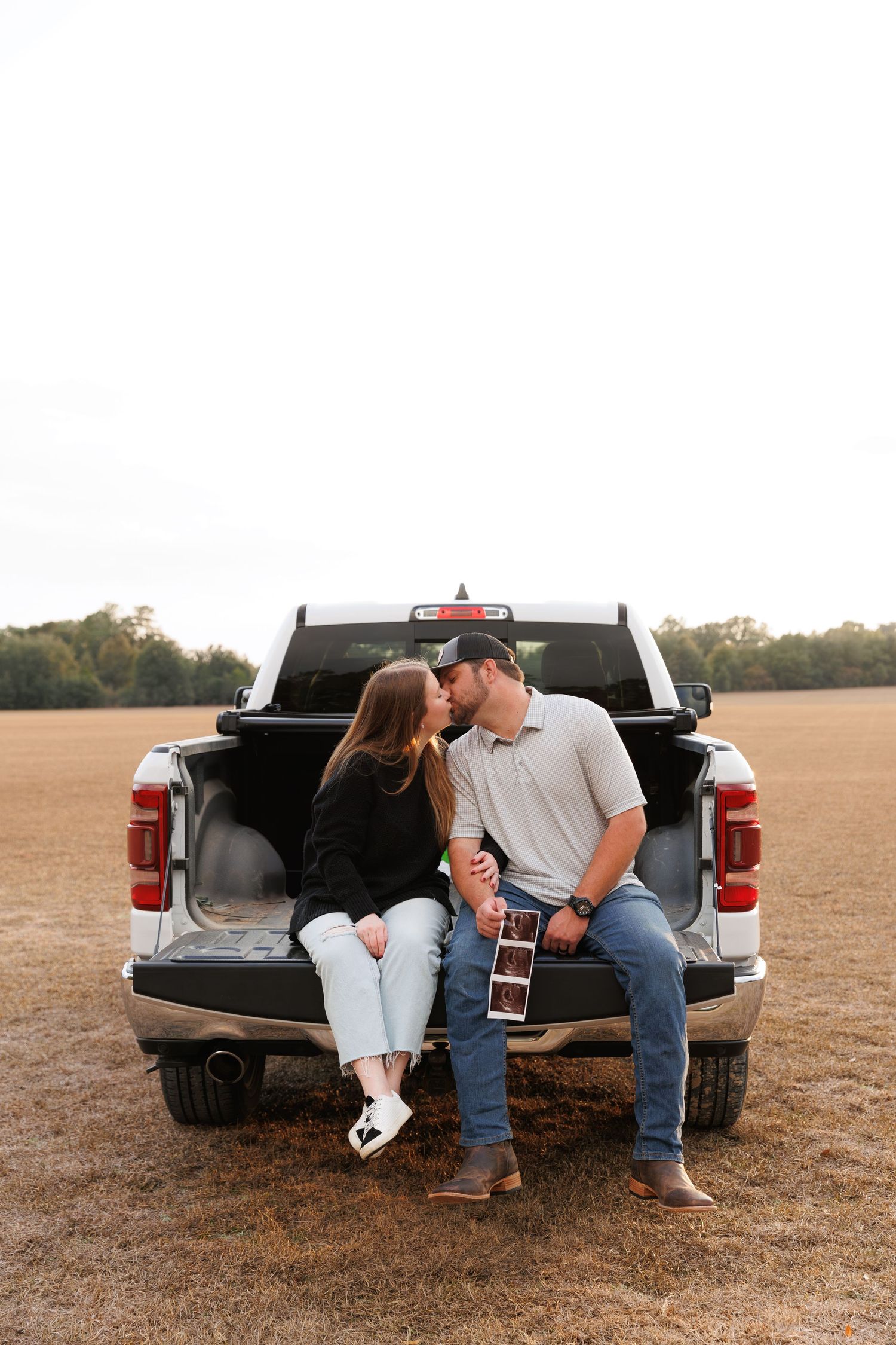 Couple sits intimately on pickup truck tailgate in rustic field during golden hour.