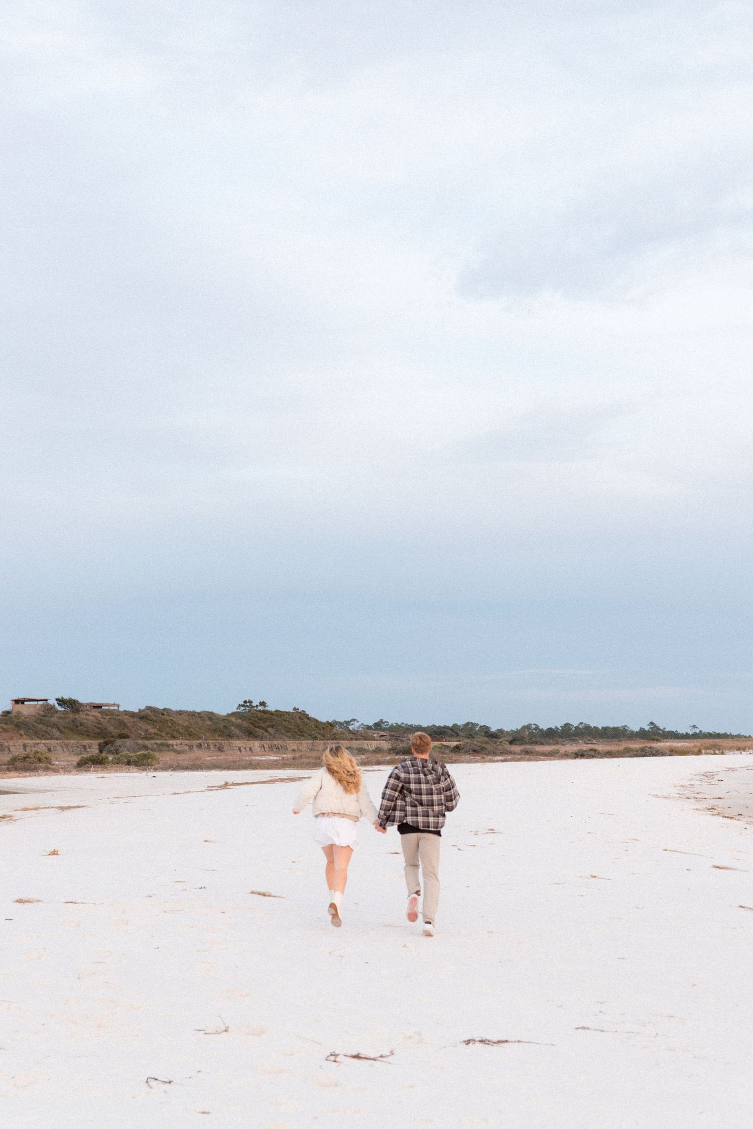 Two people walking together on a pristine white sandy beach with overcast skies and coastal vegetation.