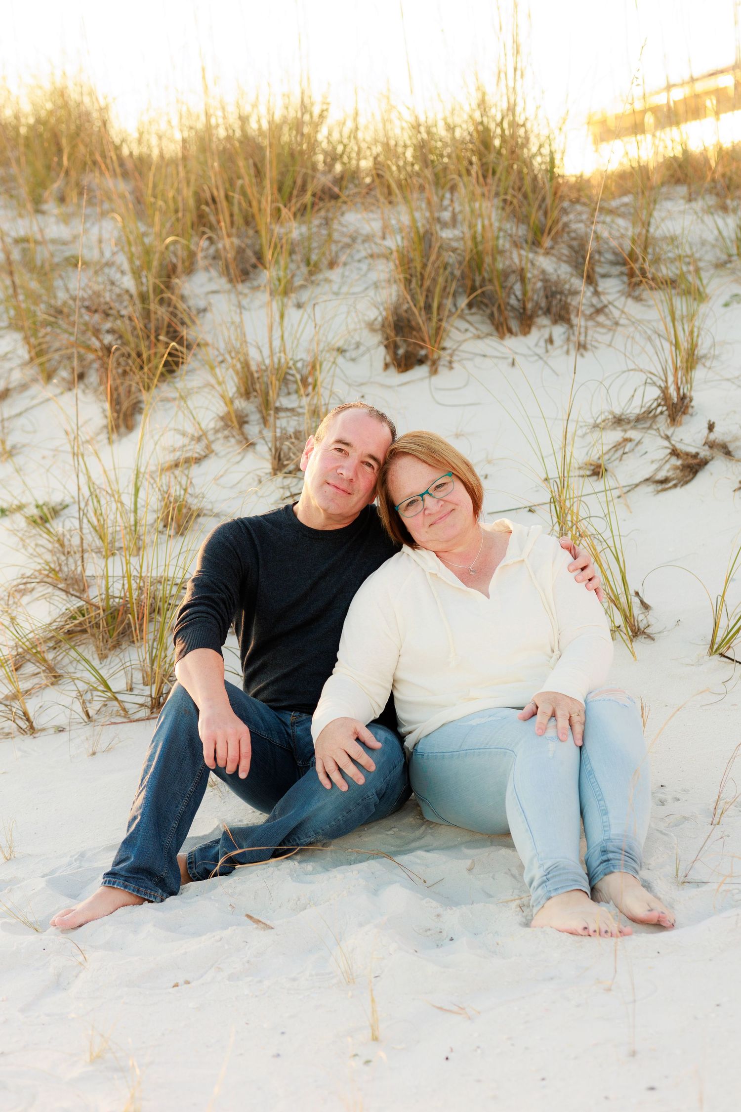A couple relaxes together on a sandy beach dune at sunset with tall grass swaying in the background.