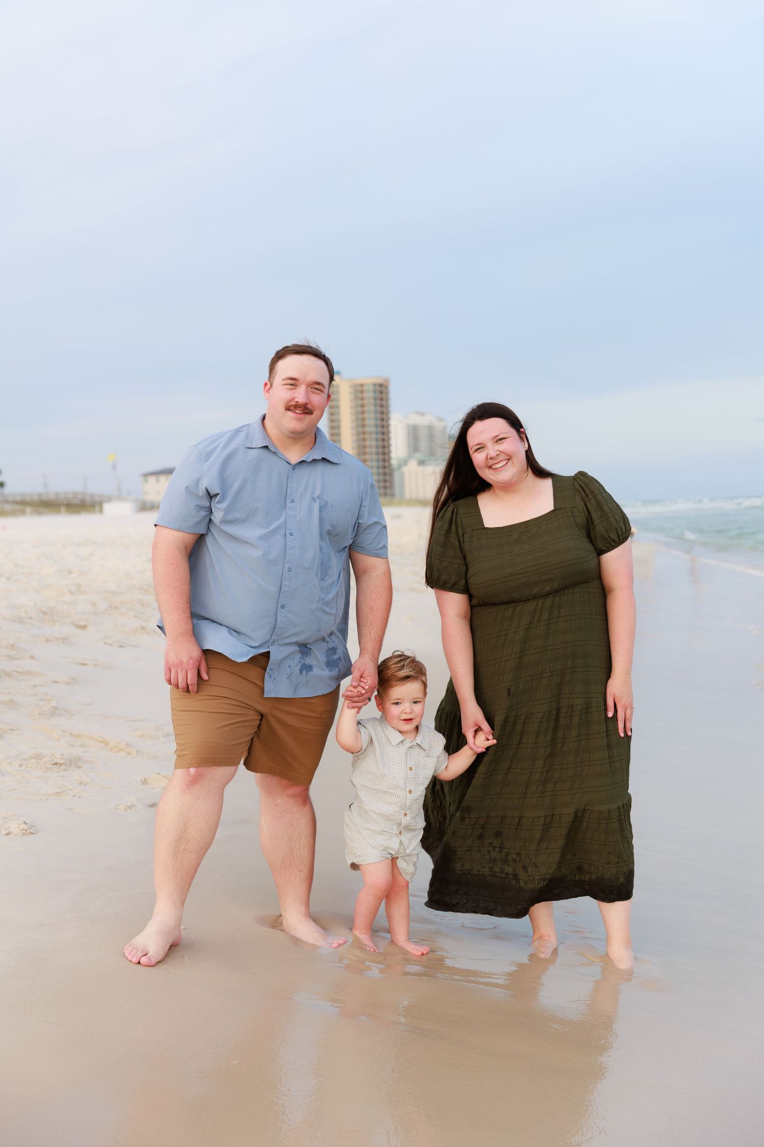 A family of three poses together on a sandy beach with coastal buildings visible in the background.