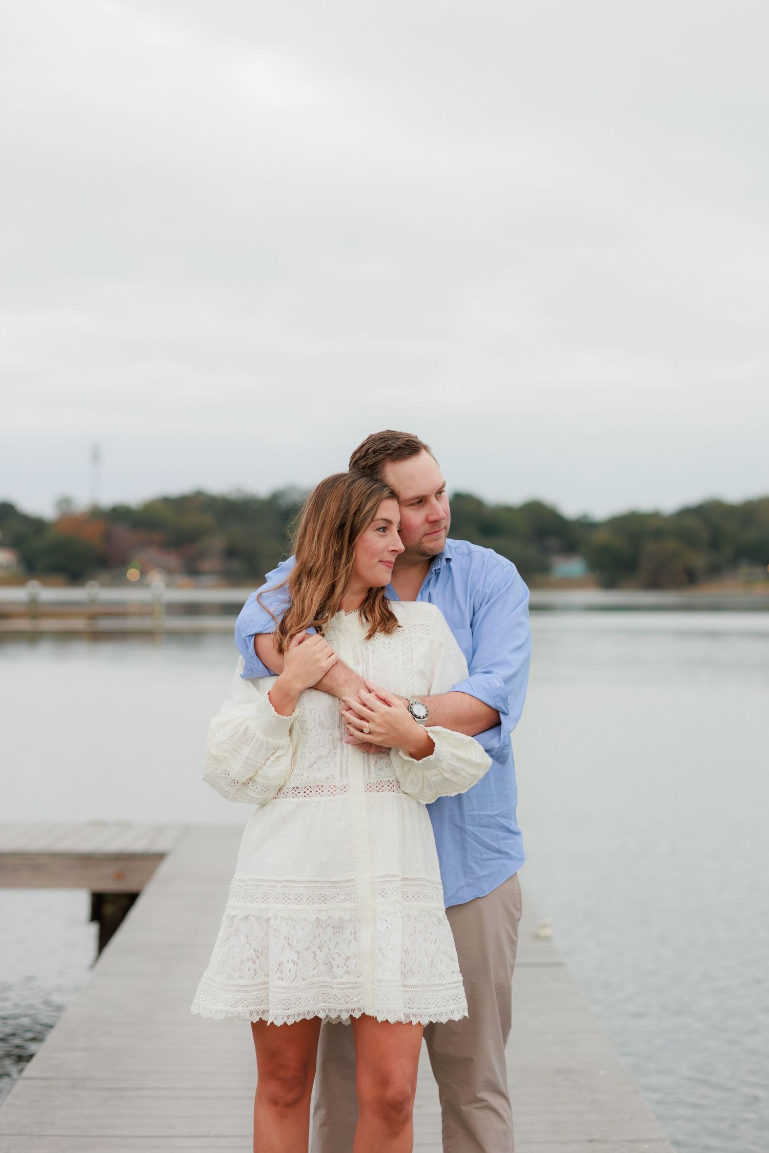 A couple embraces tenderly on a wooden dock overlooking a serene lake on an overcast day.