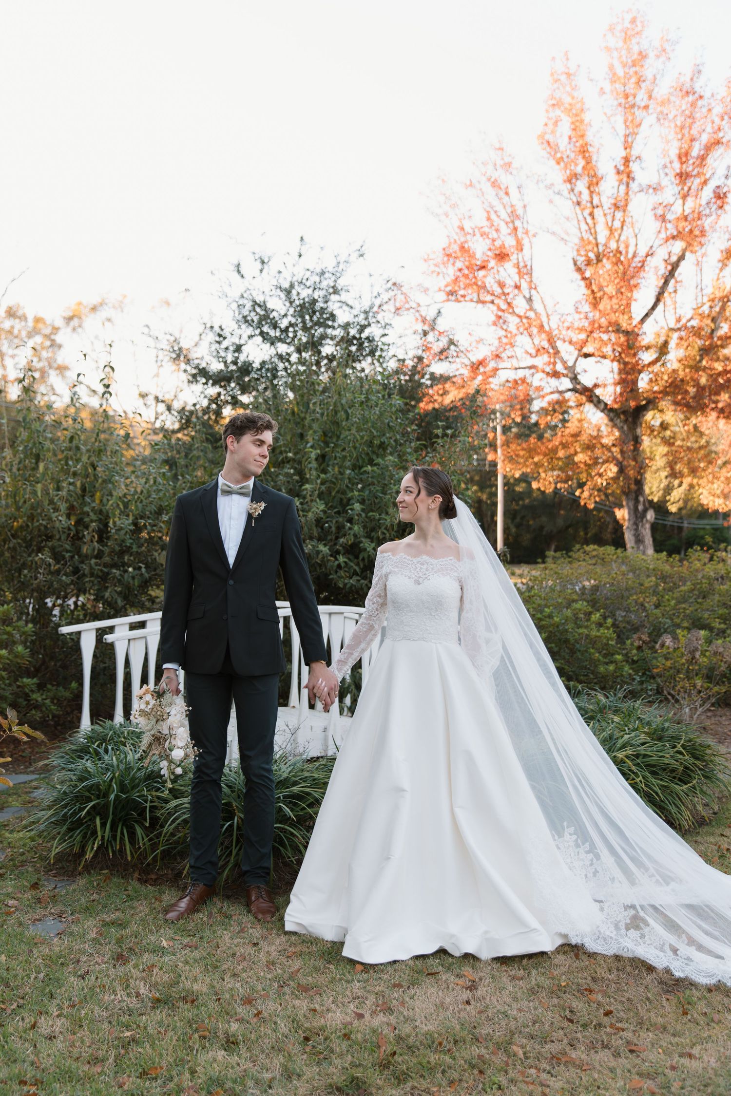 Newlywed couple walking through garden at sunset with bride wearing long-sleeved lace gown and flowing cathedral veil.