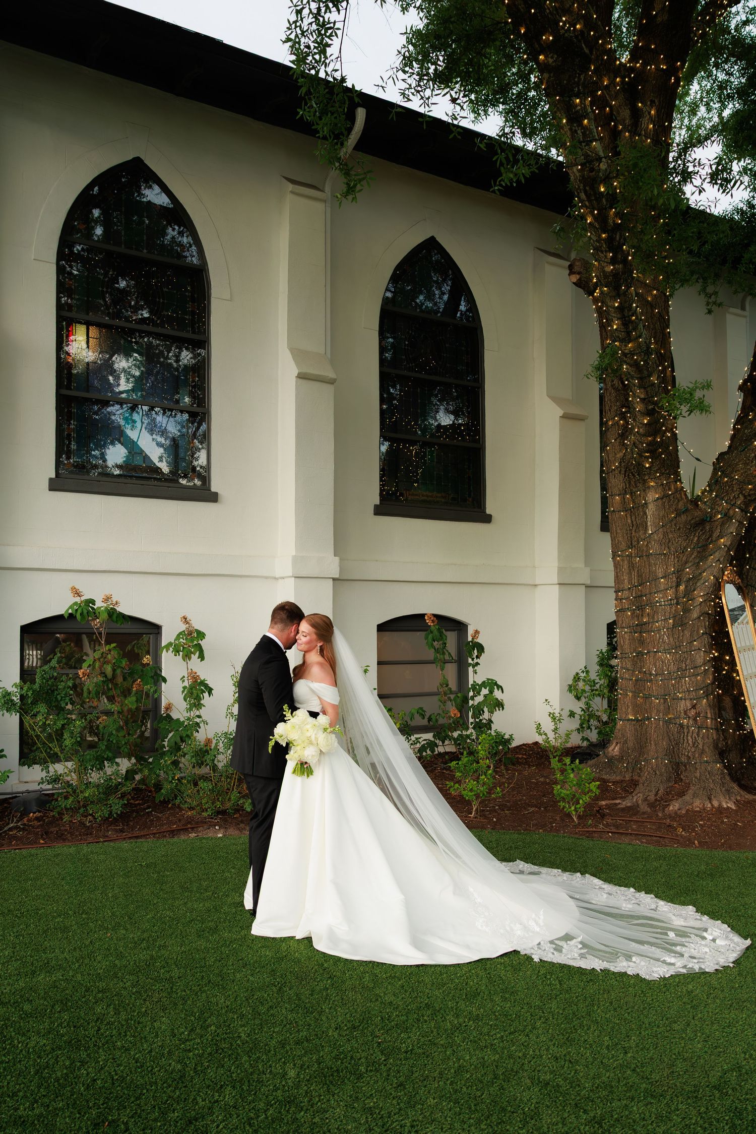 Bride and groom sharing romantic kiss outside white chapel with gothic windows and twinkling tree lights.