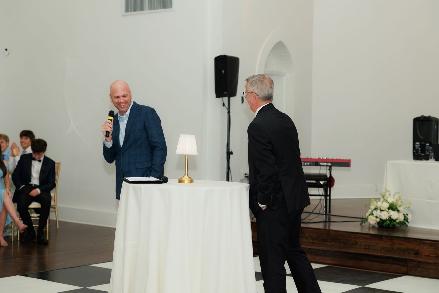 Man speaking into microphone at elegant wedding reception with white tablecloth and floral arrangements in bright venue.