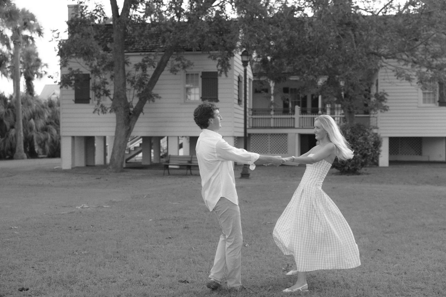 Black and white photo of a couple dancing outdoors on a lawn in front of a historic Southern home.
