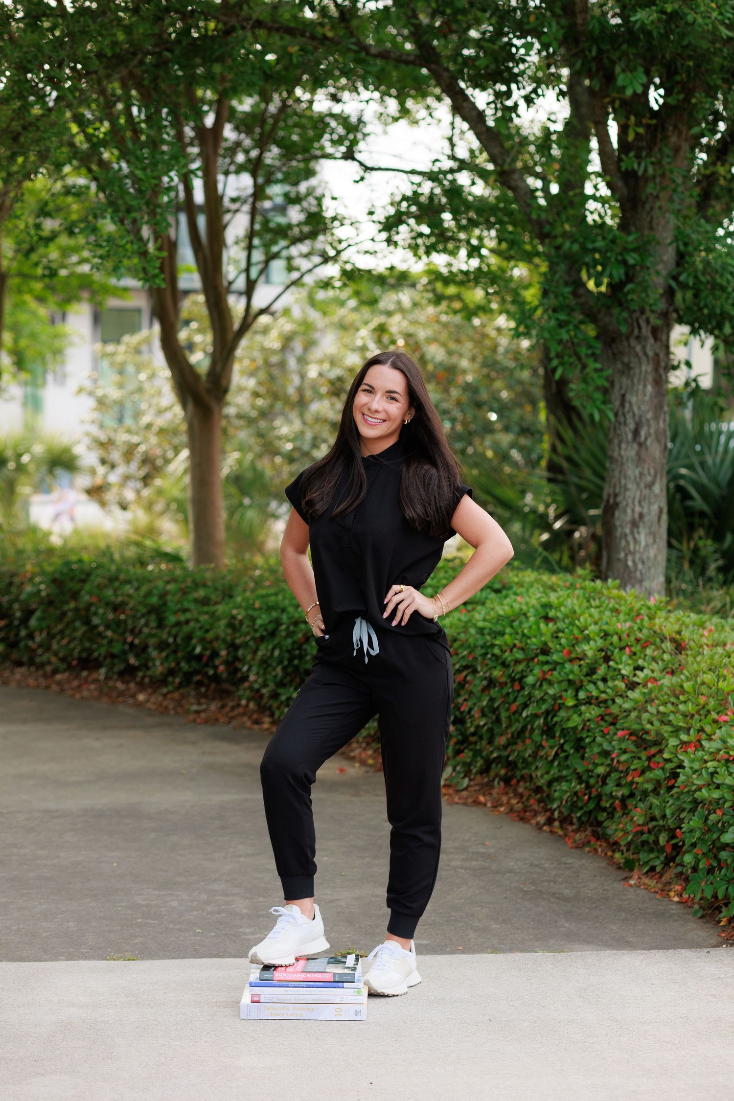 Woman in black athletic outfit standing confidently outdoors, foot on books, surrounded by lush green trees and shrubs.