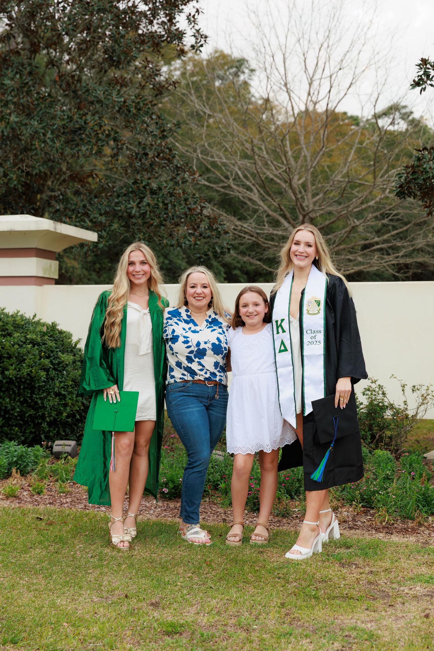 Four women pose outdoors at a graduation celebration, two in academic regalia with green and black gowns.