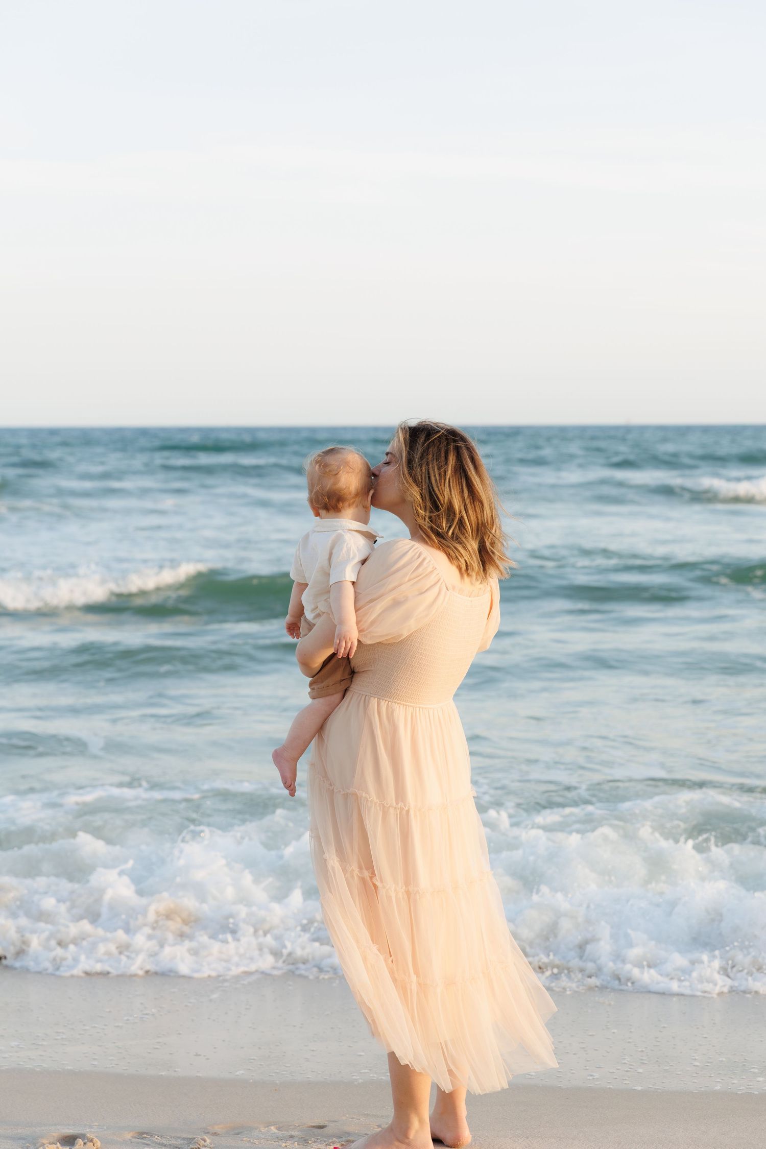 Mother in peach dress kissing baby while standing at the ocean's edge on a beach.
