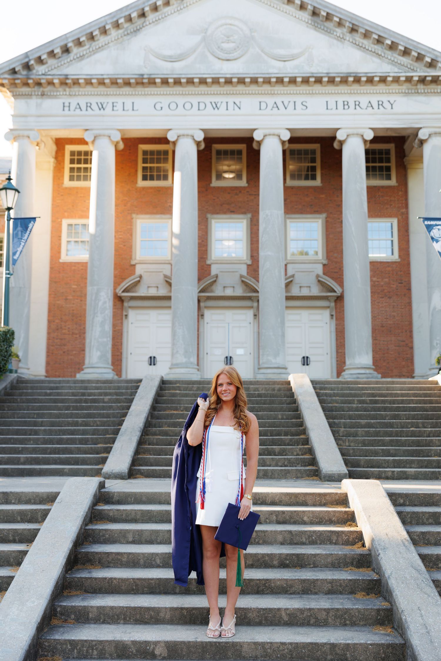 Graduate in white dress holds cap and gown on steps of Harwell Goodwin Davis Library at university.